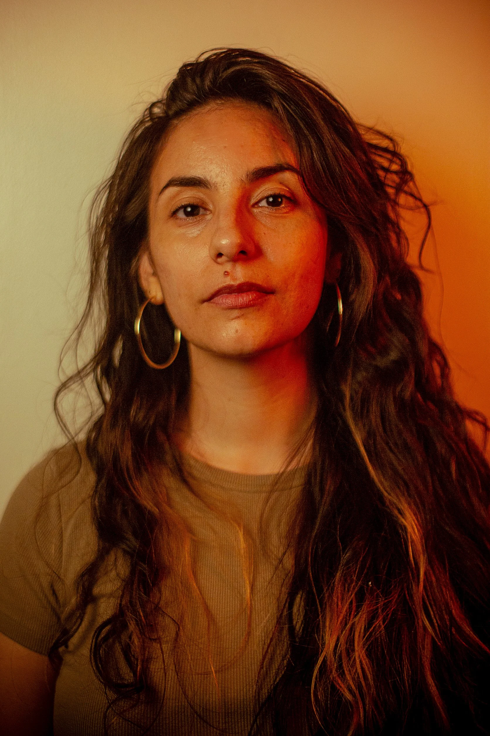 A woman with long curly hair, wearing large gold hoop earrings and a tan top, standing against a warm, gradient background.