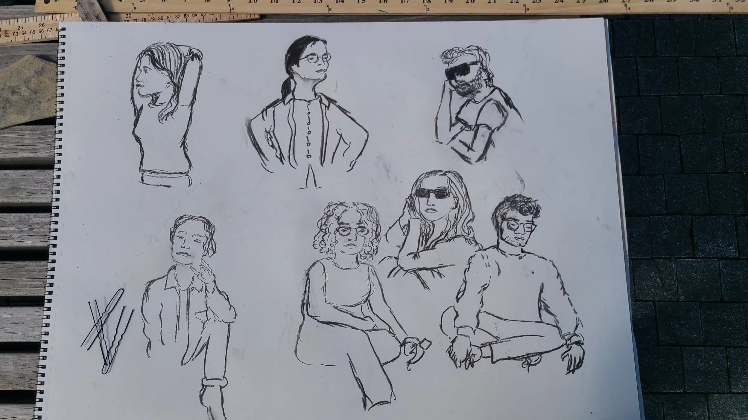 A sketchpad with various black ink drawings of people, including women and men, in different poses and expressions, laid on a wooden surface with some drawing tools nearby.