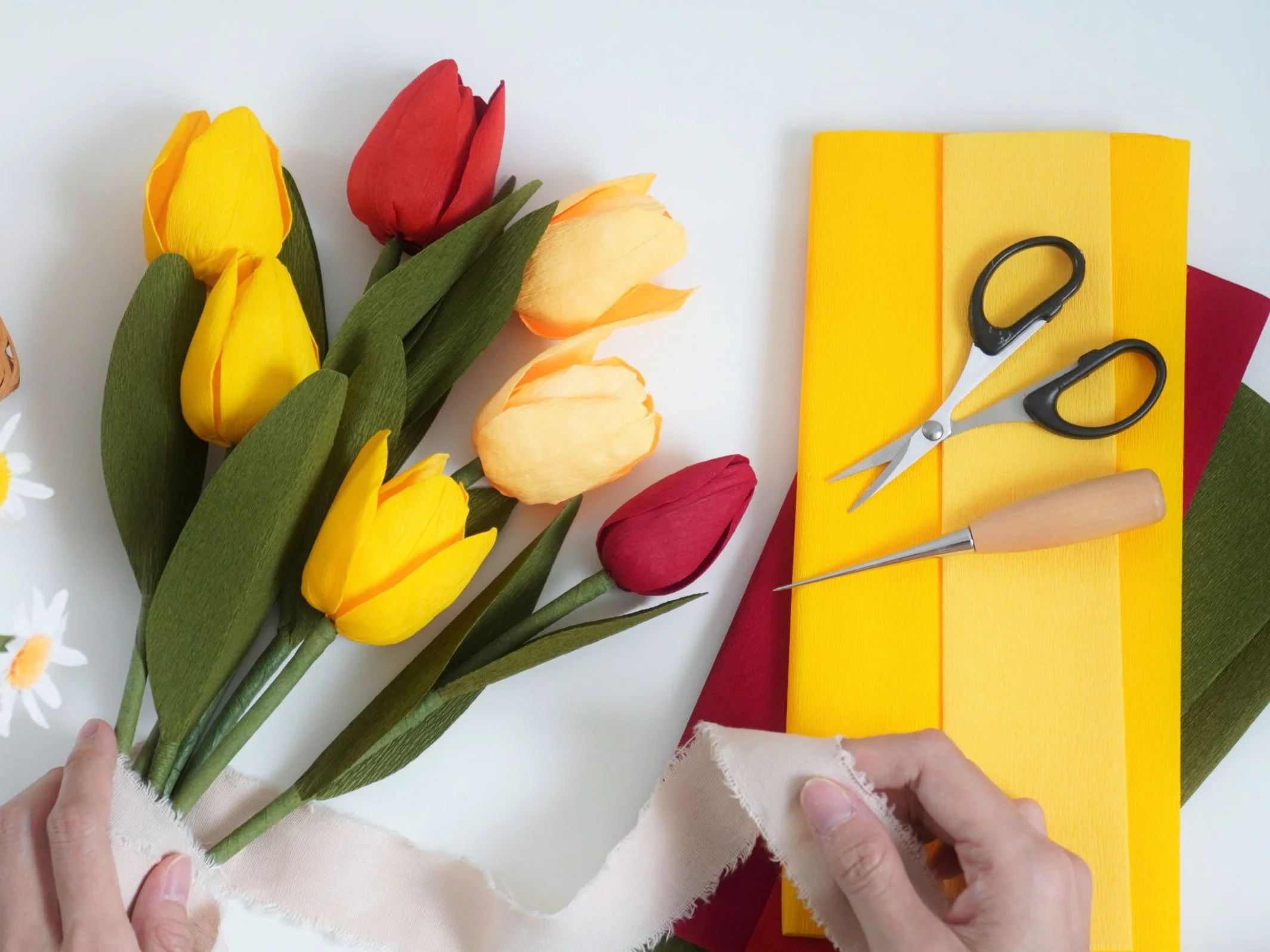 Jasmine Sing, paper artist arranging handmade paper tulip flowers on a white surface with yellow and red foam sheets and scissors nearby.