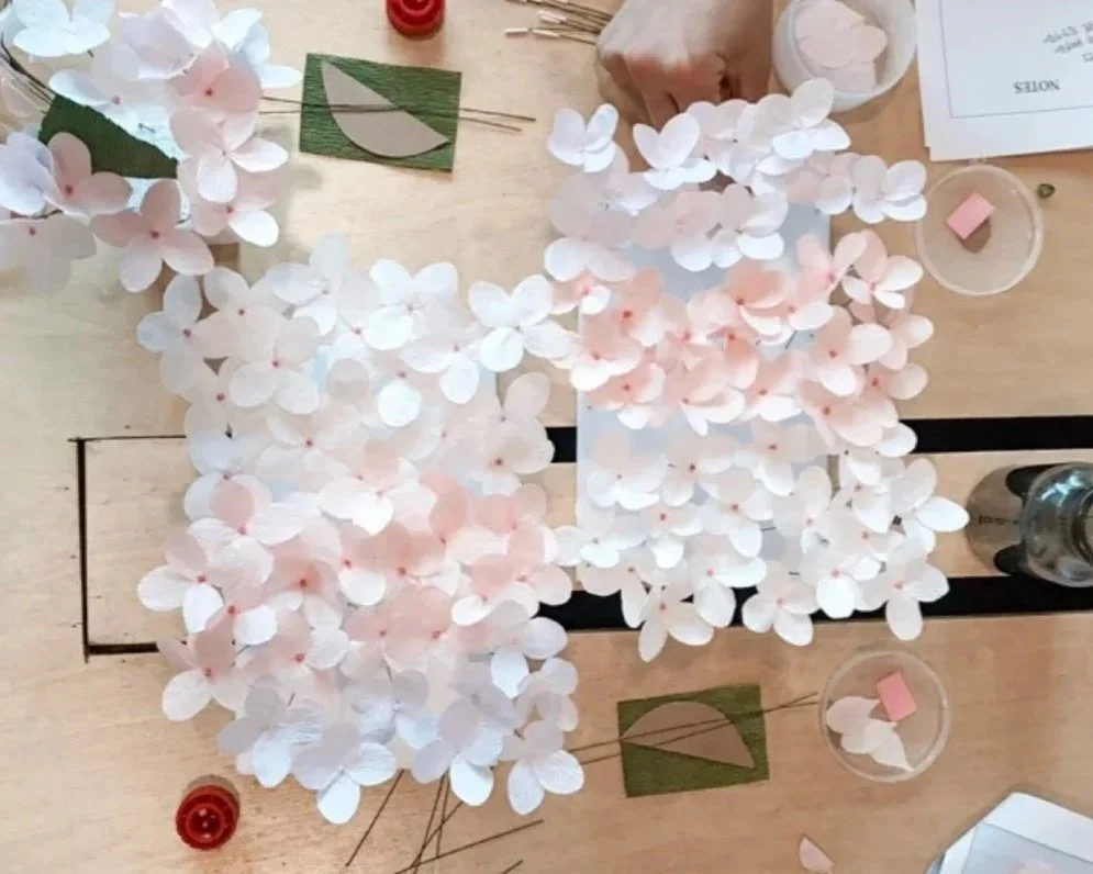 Crafting table with pink and white paper hydrangea flowers, green leaves.