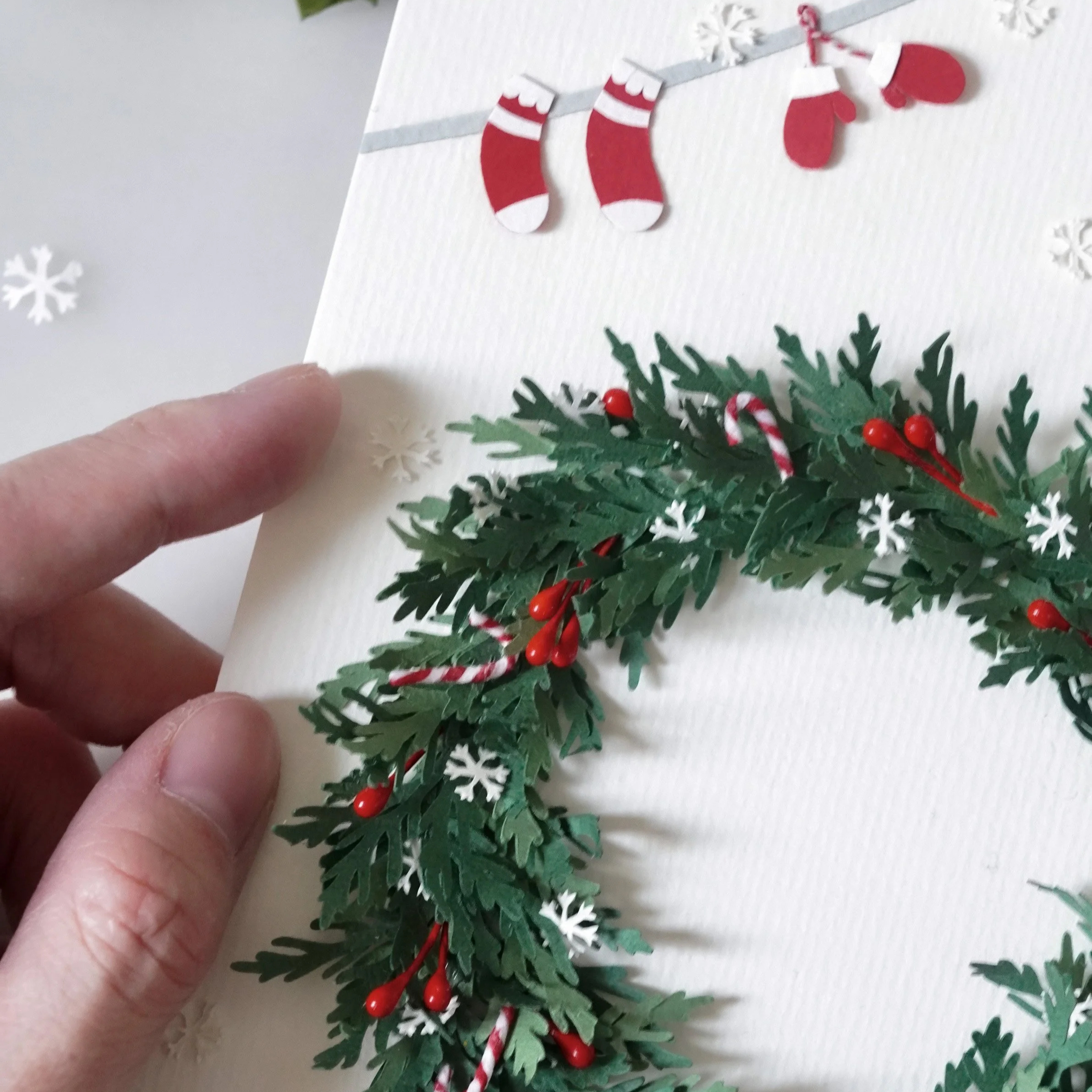 Close-up of a holiday card featuring red Christmas stockings, green holly leaves, red berries, and white snowflakes,  handmade by Jasmine Sing, paper artist from Merremade for Christmas season gifting.