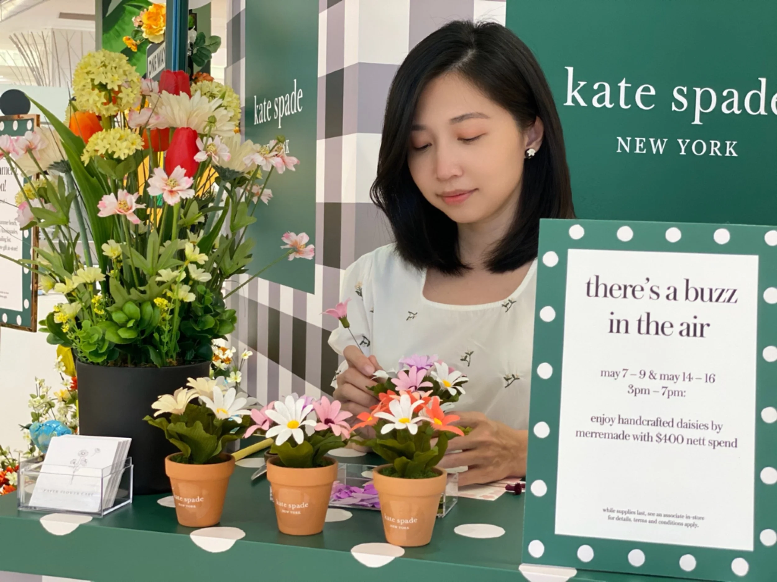 Jasmine Sing, paper artist arranging colorful paper daisies in small terracotta pots at a display booth for Kate Spade New York in Singapore. Behind her, there is a green sign with white text promoting handcrafted daisies for sale.