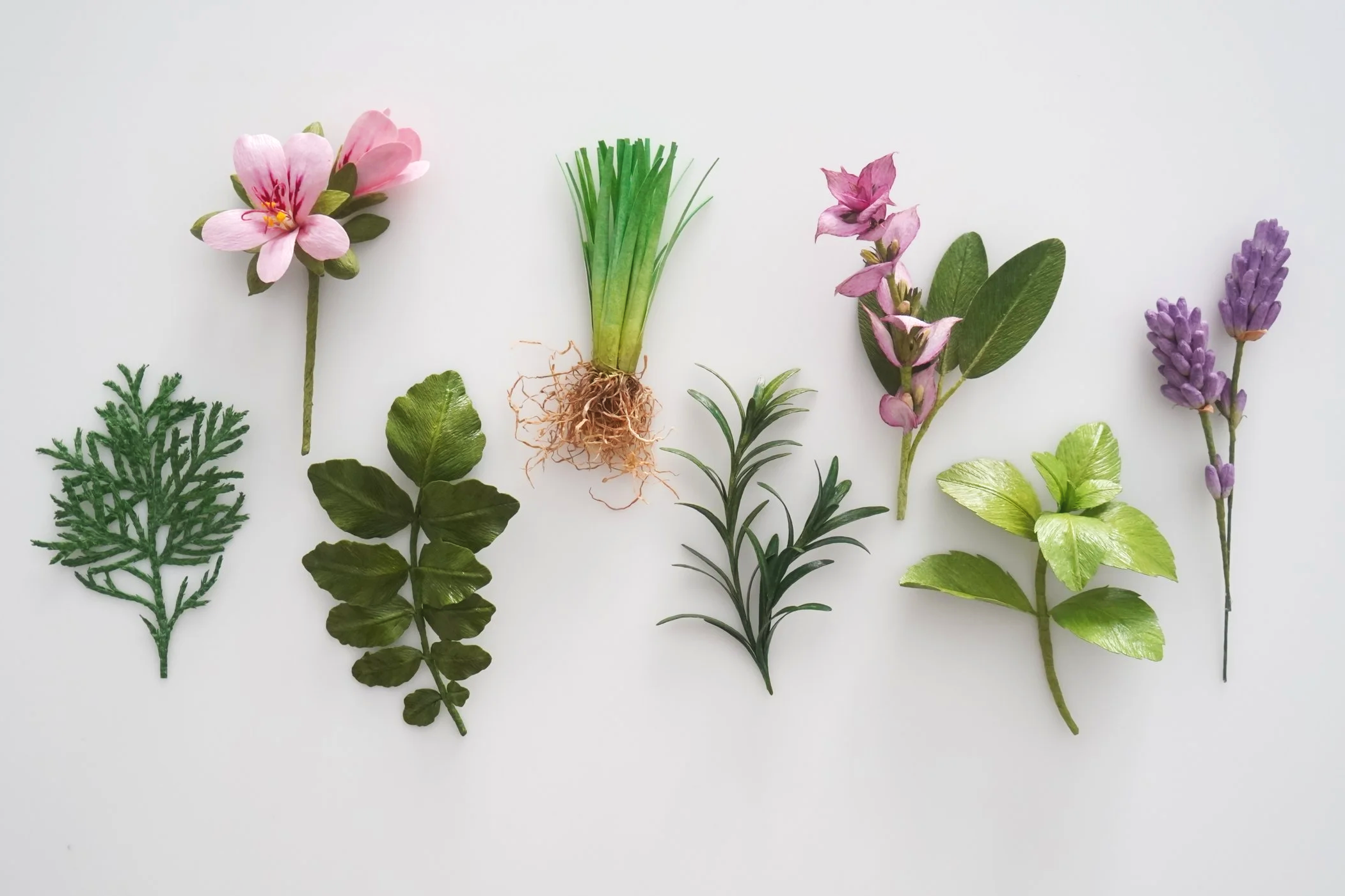 Array of realistic paper herbs, flowers and leaves on a white background, including pink blossoms, purple lavender, and green foliage.