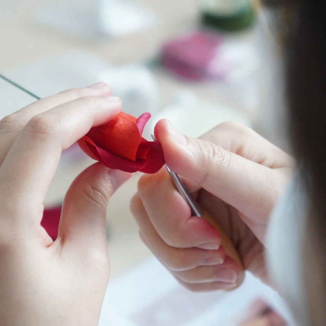 Merremade student is carefully crafting a very detailed red and pink paper flower, using a small tool to shape the petals.