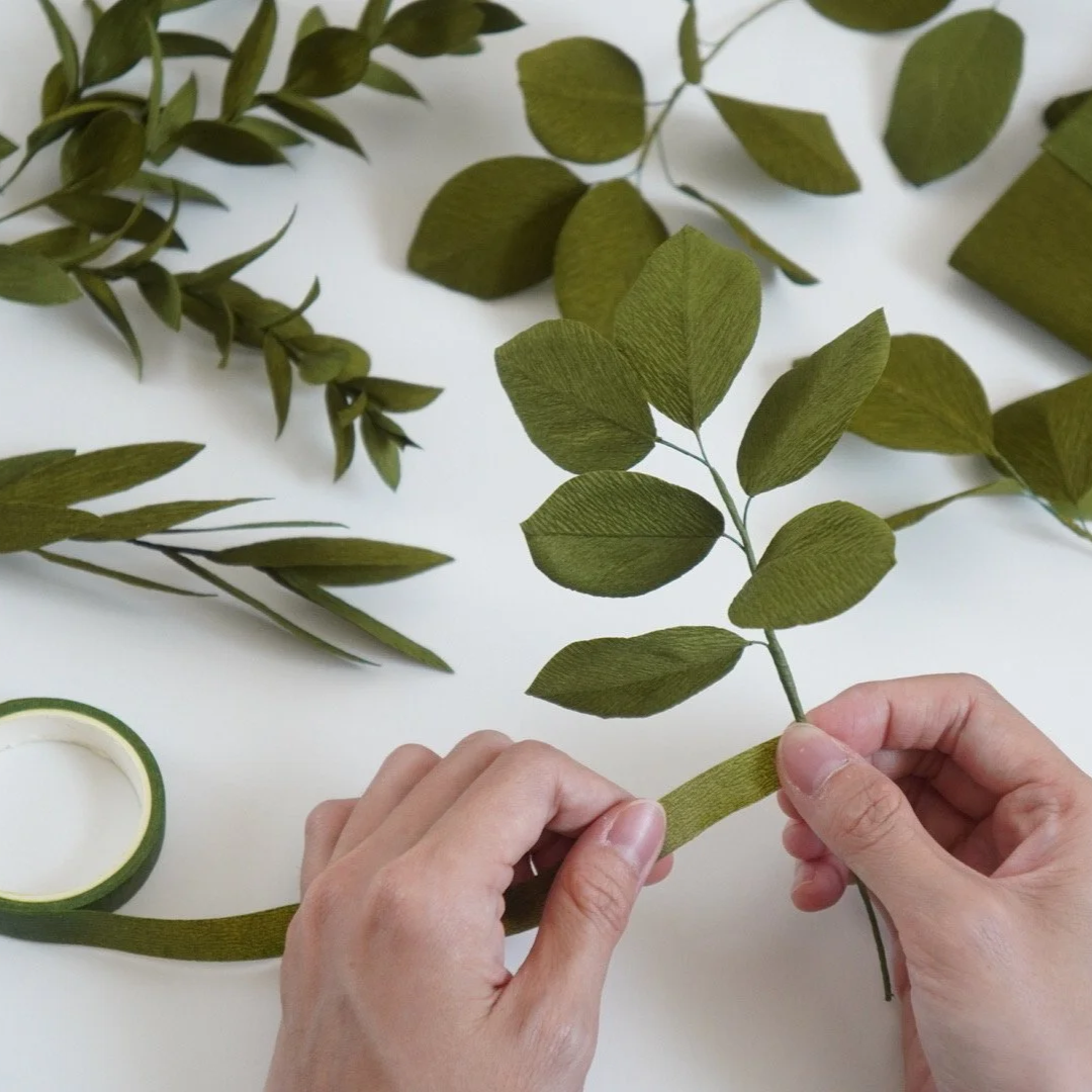 Jasmine Sing, paper artist arranging handmade paper green leaves and vines on a white surface with a roll of green floral tape.