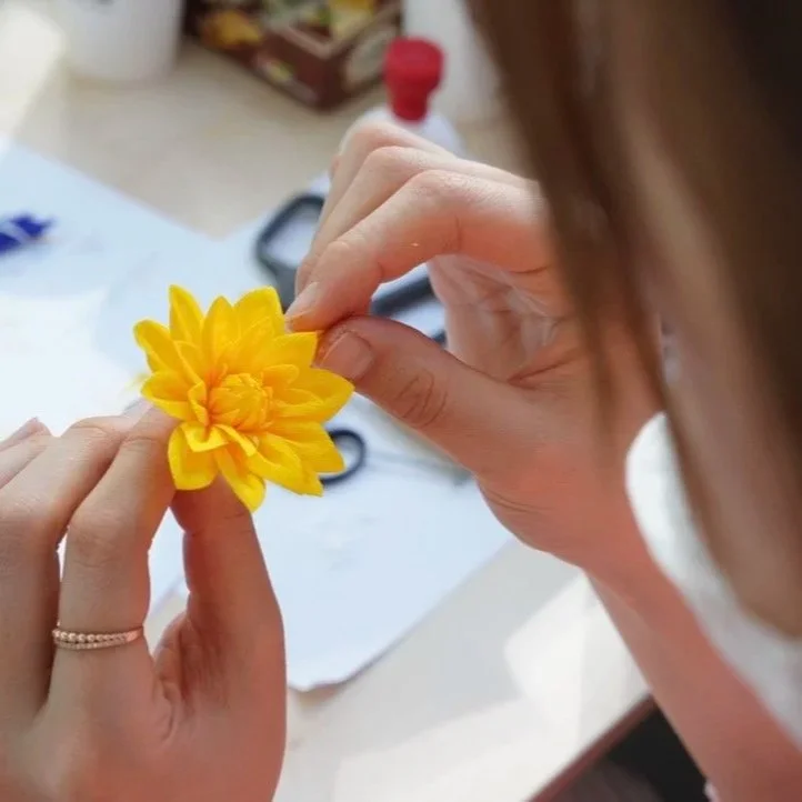 Merremade student holding a handmade yellow paper flower with scissors nearby on a table during in-person workshop in Seoul, Korea.