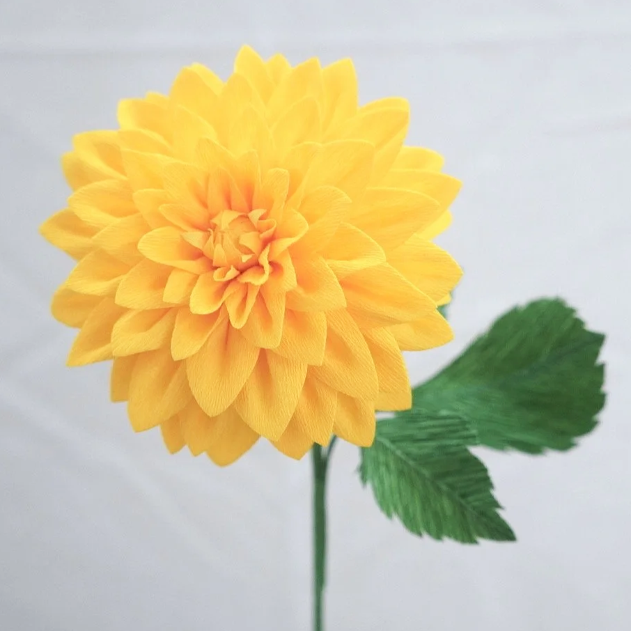 Yellow artificial flower with layered petals and two green leaves on a stem, against a plain white background.