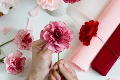 Artist arranging handmade pink and red paper flowers on a table with various rolls of pink and red paper.
