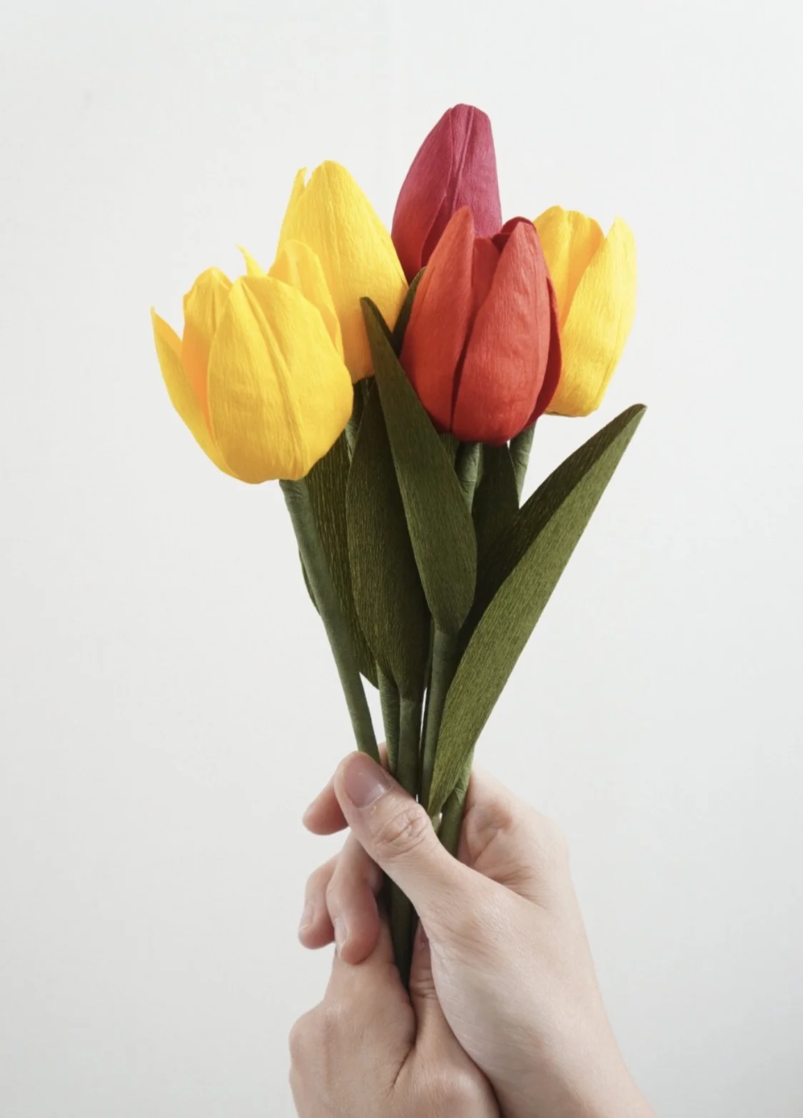 Hand holding a bouquet of handmade yellow and red paper tulips with green leaves against a plain white background.