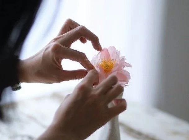 Hands arranging a handmade pink paper flower in a white vase near a window.