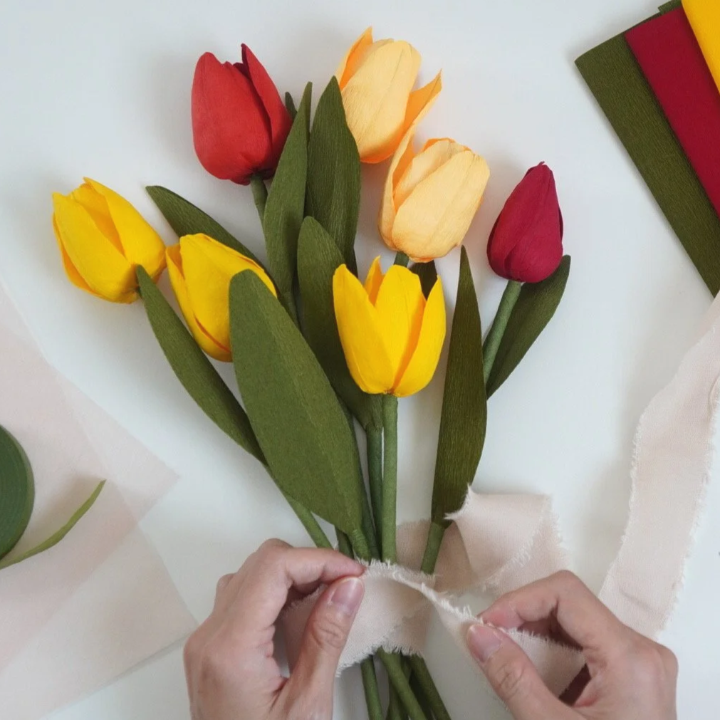 Jasmine Sing, paper artist tying a ribbon around a bouquet of handmade tulip flowers in red, yellow, and peach colors.