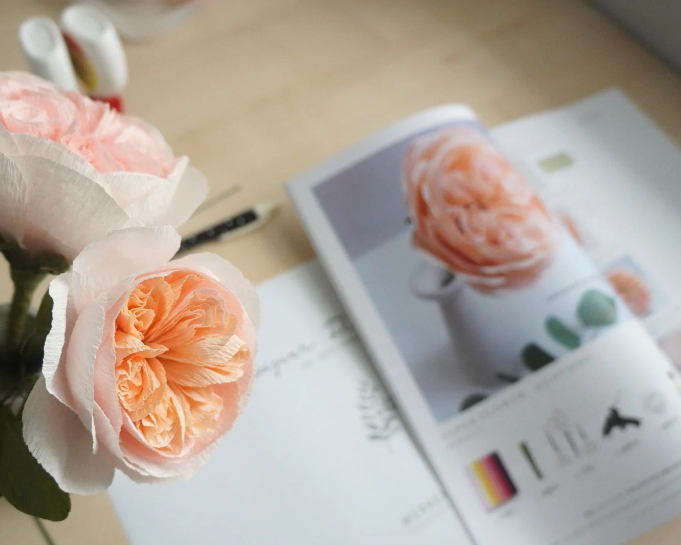 Close-up of handmade paper peach-colored Juliet roses on a desk with an open magazine and some supplies in the background.