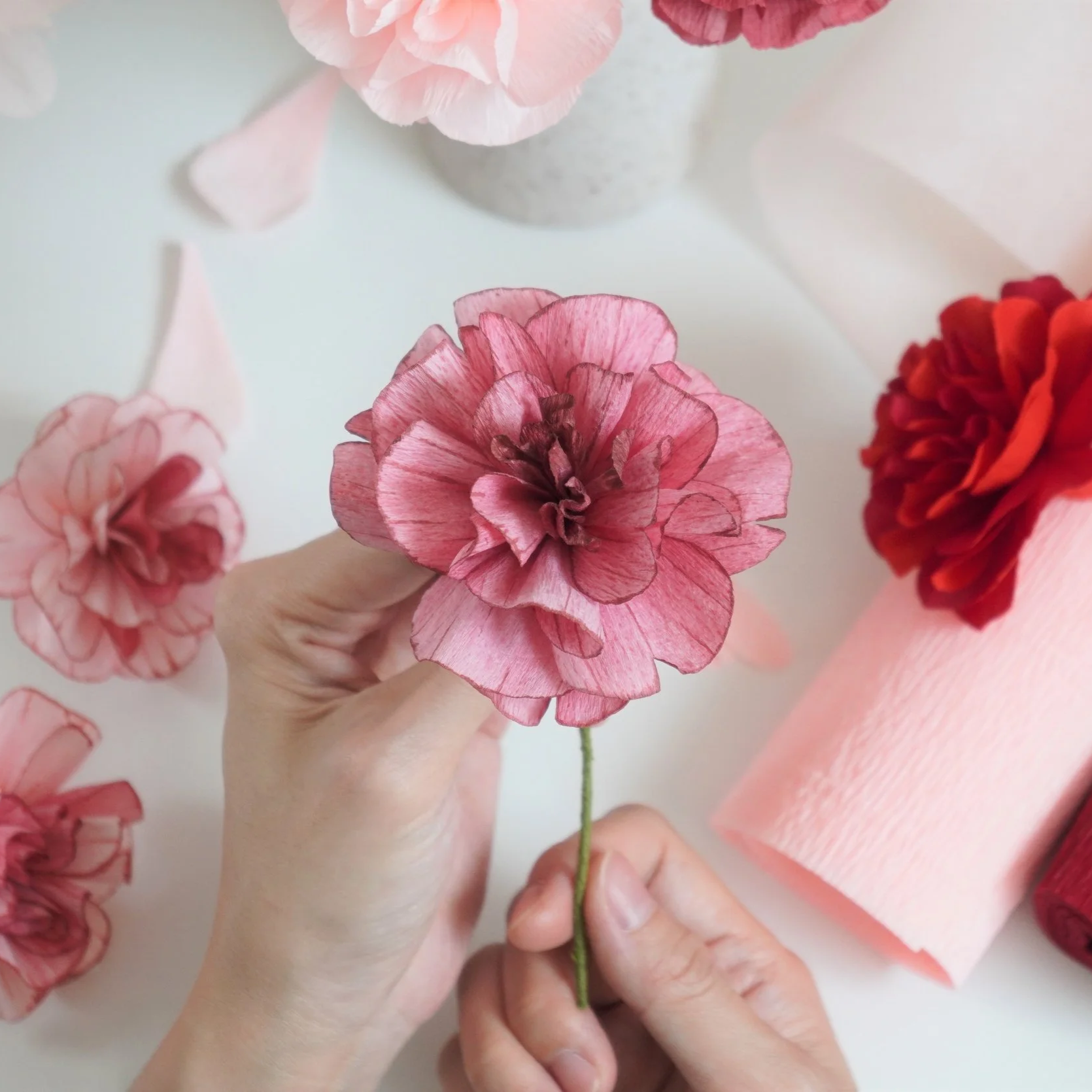 Close-up of hands holding a pink and red paper flower with a green stem, surrounded by other paper flowers and pink crepe paper decorations on a light background.