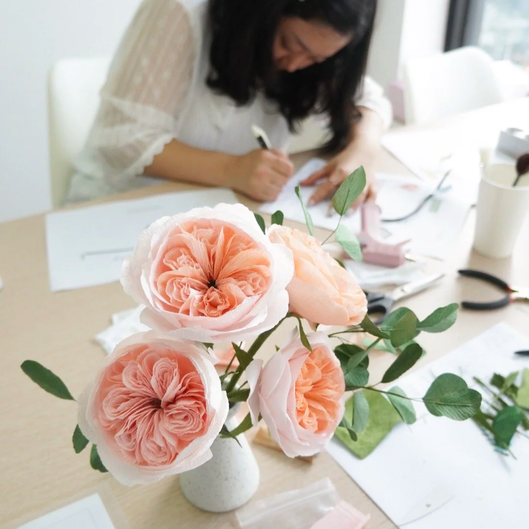 A close-up of a bouquet of handmade peach-colored paper Juliet roses with lush petals and green leaves on a light-colored table during a in-person workshop in Seoul Korea.
