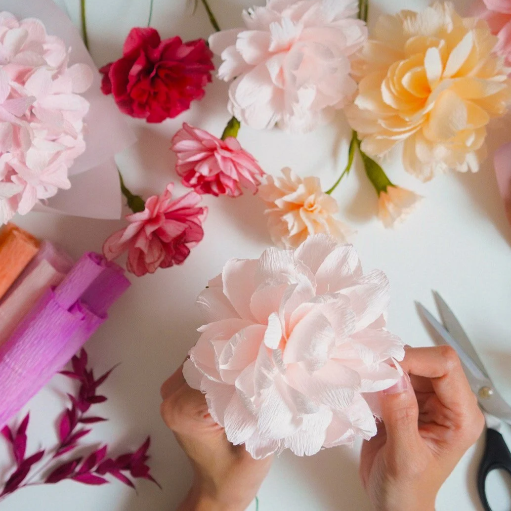 Jasmine Sing, paper artist arranging pink and cream paper flowers on a white surface, with scissors nearby.