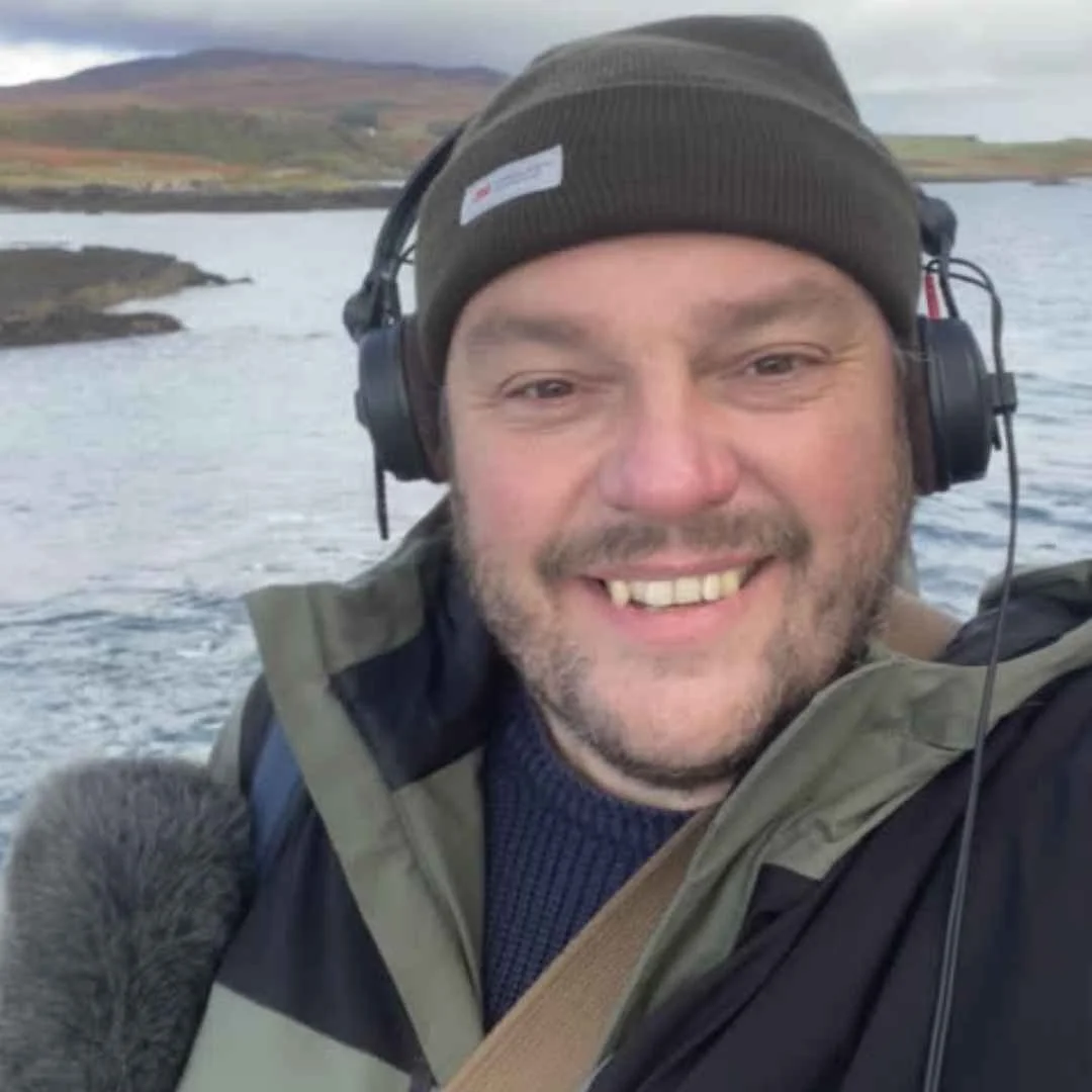 A man smiling wearing a beanie and headphones outdoors near a body of water with hills in the background.