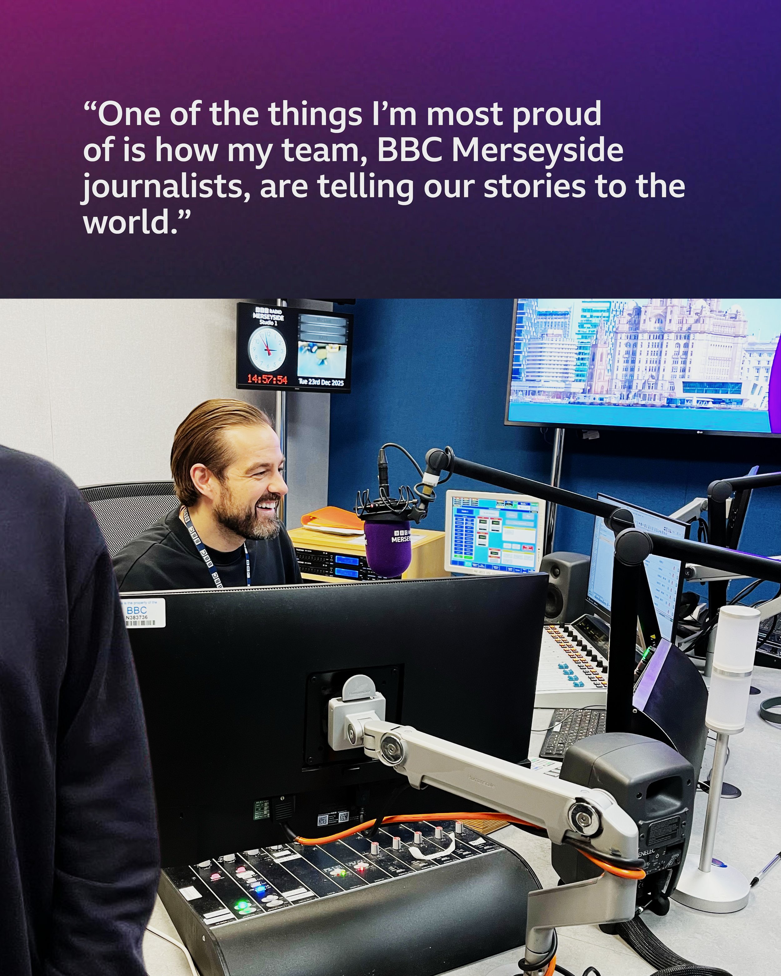 A studio radio broadcast room with a man smiling and wearing a black shirt, surrounded by electronic equipment, monitors, microphones, and a large screen displaying a cityscape.