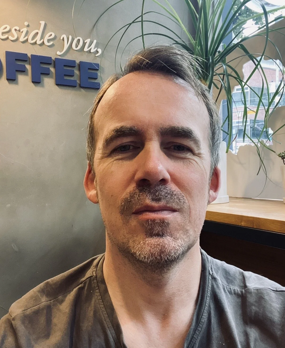 A man taking a selfie in a coffee shop with a sign on the wall that says 'beside you, FFFEE', and a large plant in the background near a window