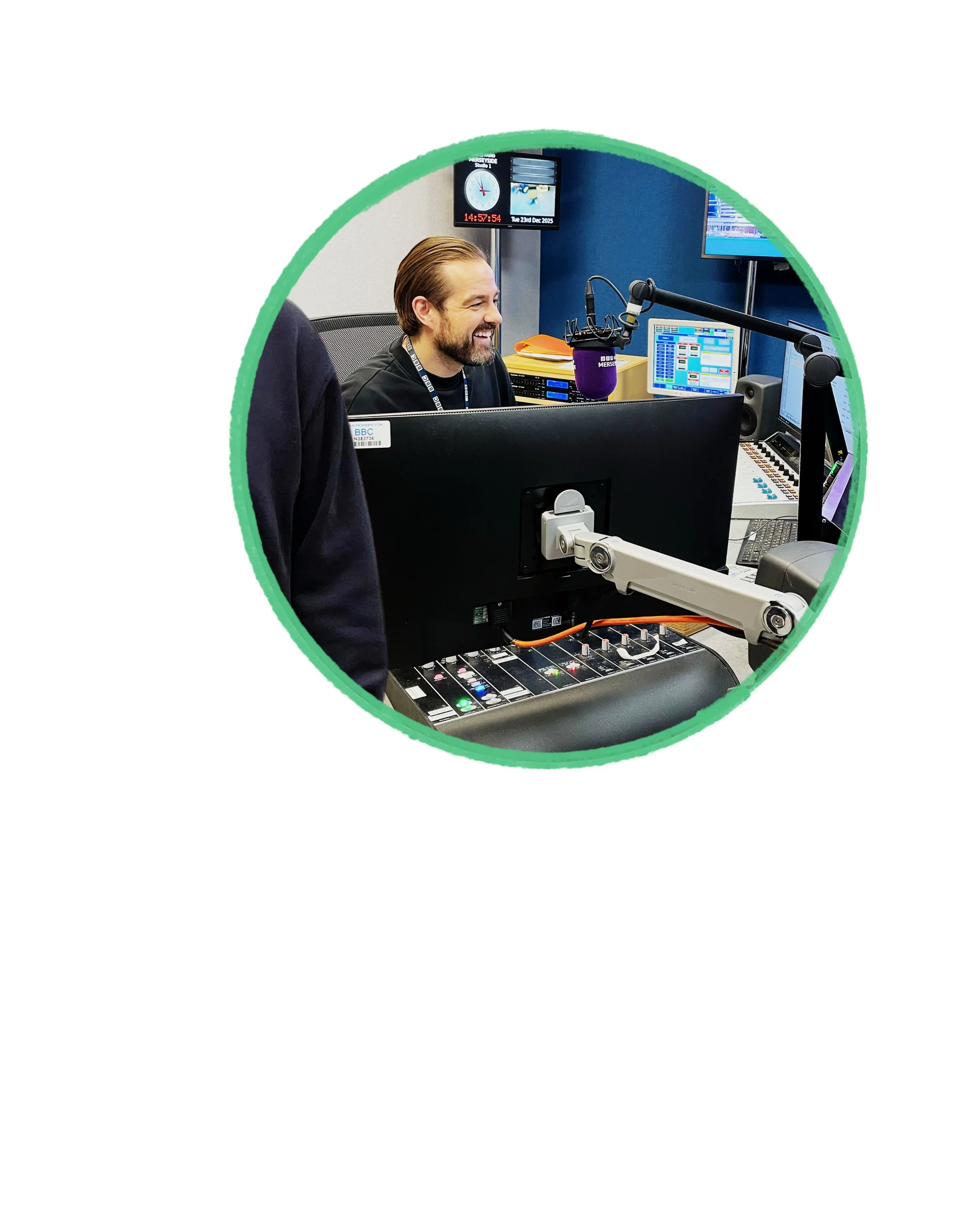 Radio host smiling in a recording studio with microphones, computer screens, and control panels.