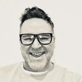 A smiling man with short dark hair, wearing glasses, in front of a plain background.