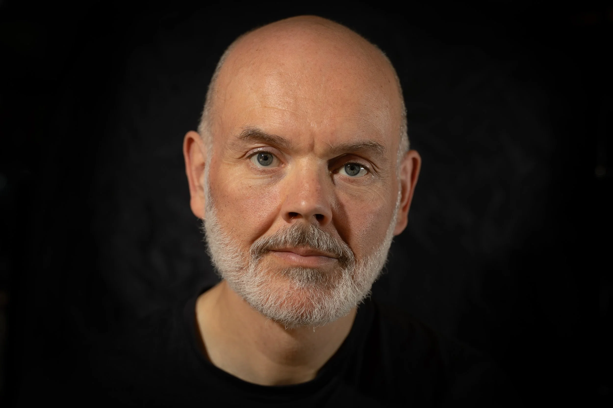 Close-up of a bald man with gray beard and blue eyes, wearing a black shirt, against a black background.