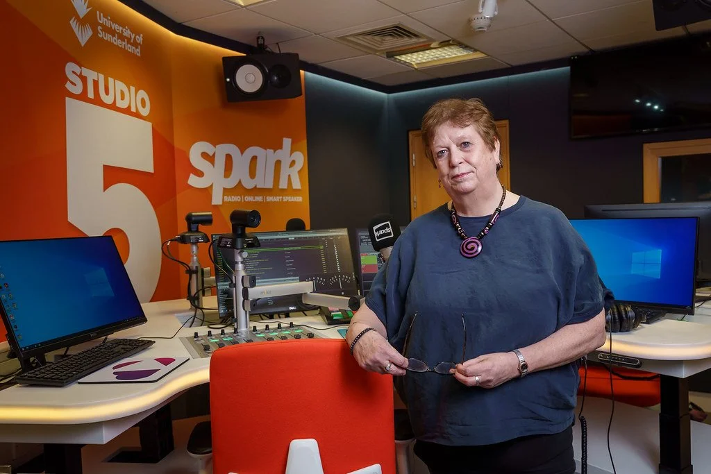 A woman standing in a radio studio with professional audio equipment, multiple monitors, and a large orange wall with the text 'Studio 5 Spark' and 'University of Sunderland' logo.