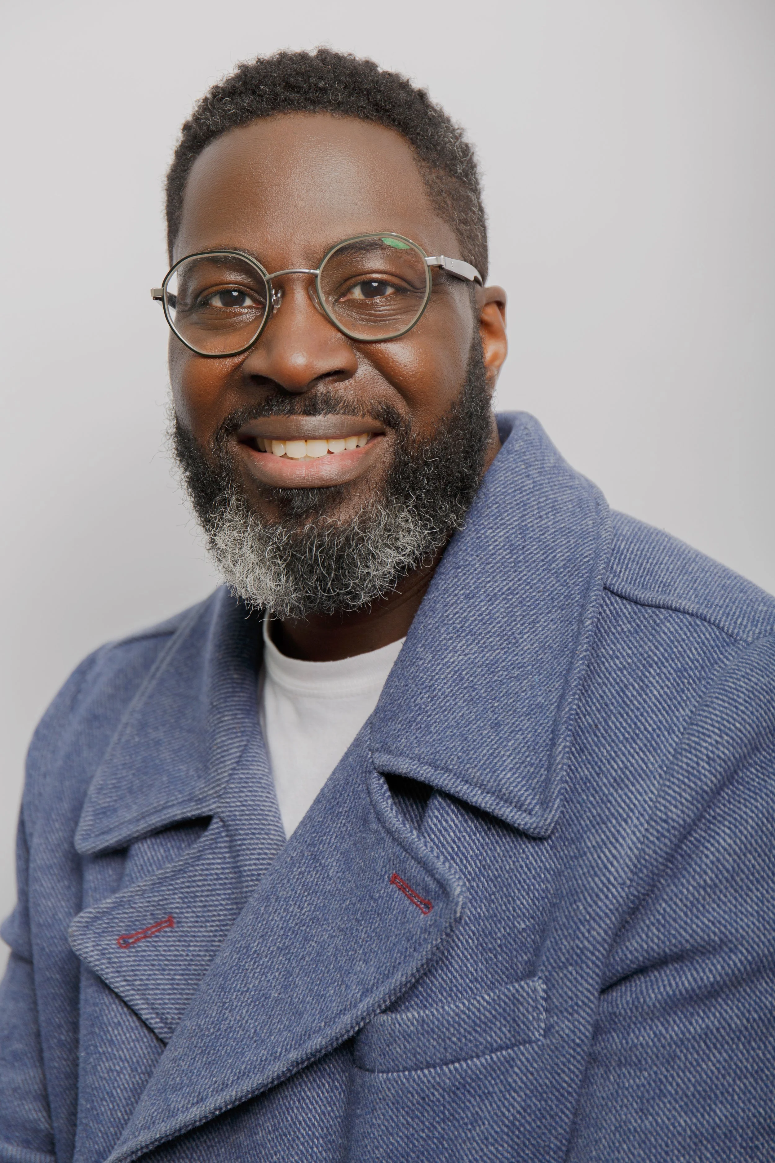 A smiling man wearing round glasses, a gray herringbone blazer, and a white shirt, with short curly hair and a beard, against a neutral light background.