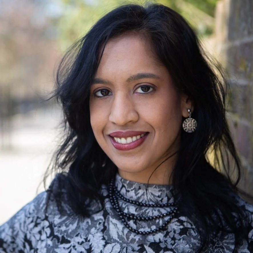 A portrait of a woman with black hair, wearing patterned clothing and jewelry, smiling outdoors near a brick wall.