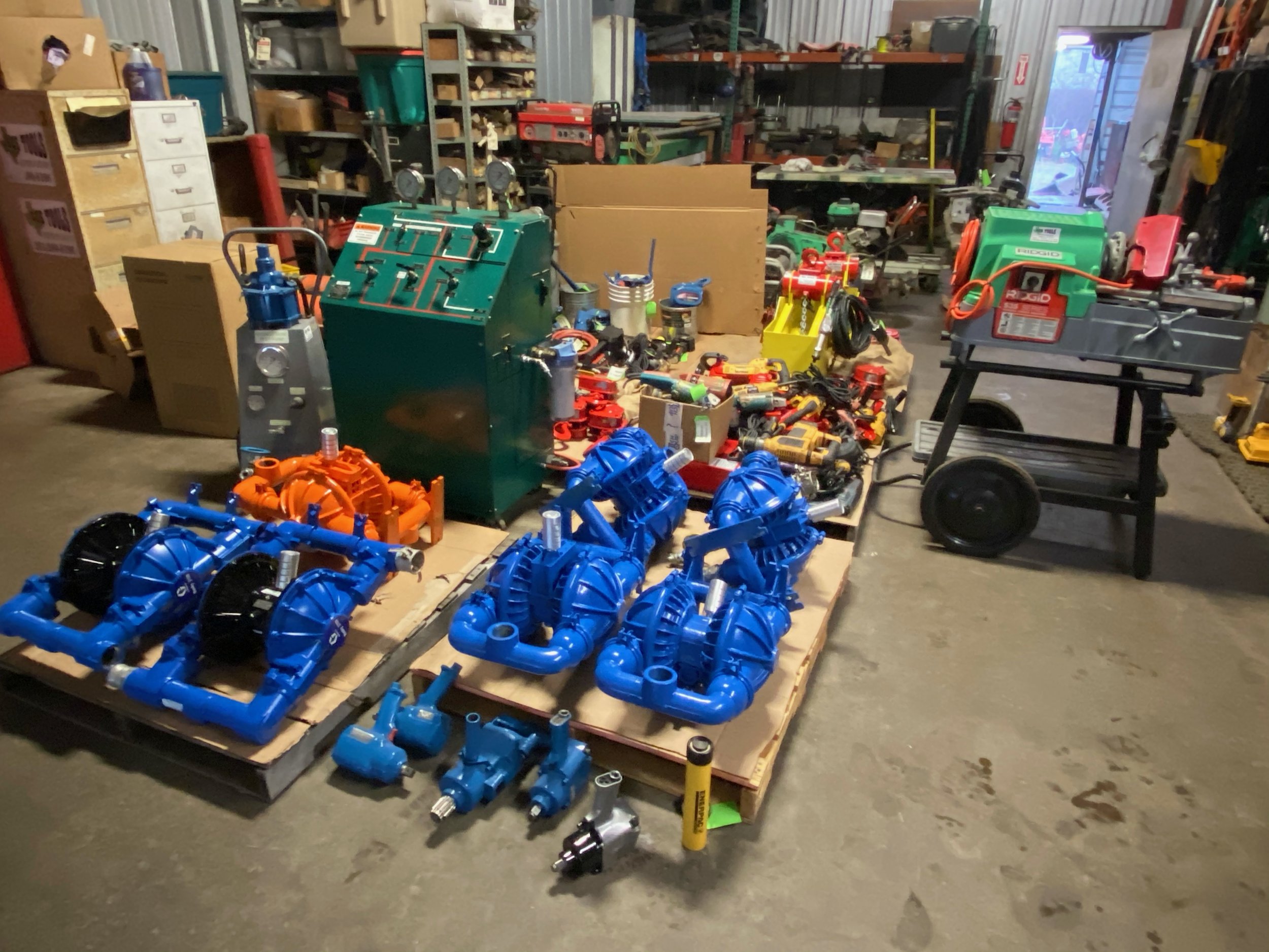 A cluttered workshop with various tools, parts, and machinery. Blue plastic pipes and black fittings are on a wooden pallet in the foreground. Shelves and workbenches are filled with equipment, boxes, and supplies in the background.
