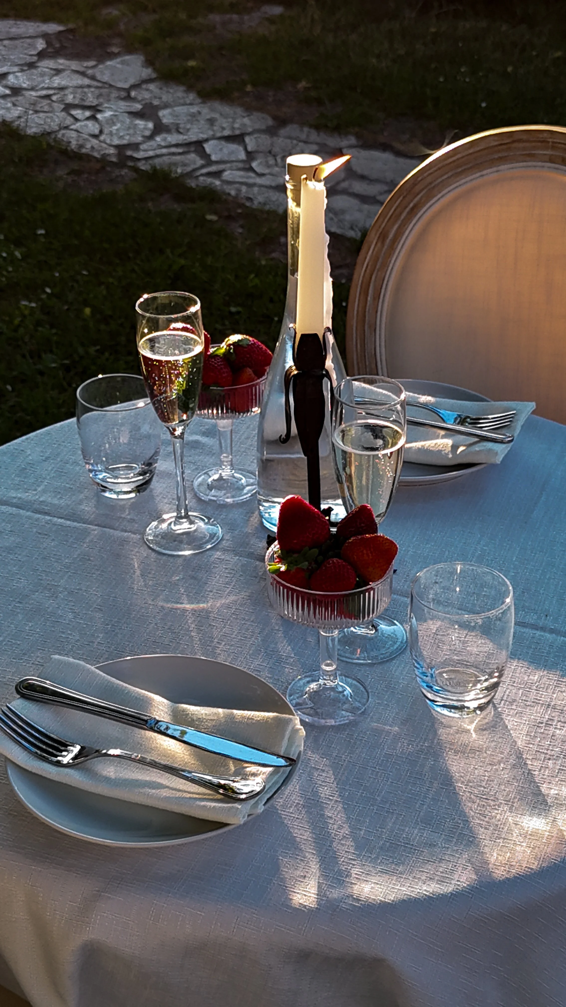 Tavolo apparecchiato con bicchieri di vino, fragole in ciotole di vetro, piatto con posate, candela accesa, e una sedia in background, in un ambiente all'aperto serale.