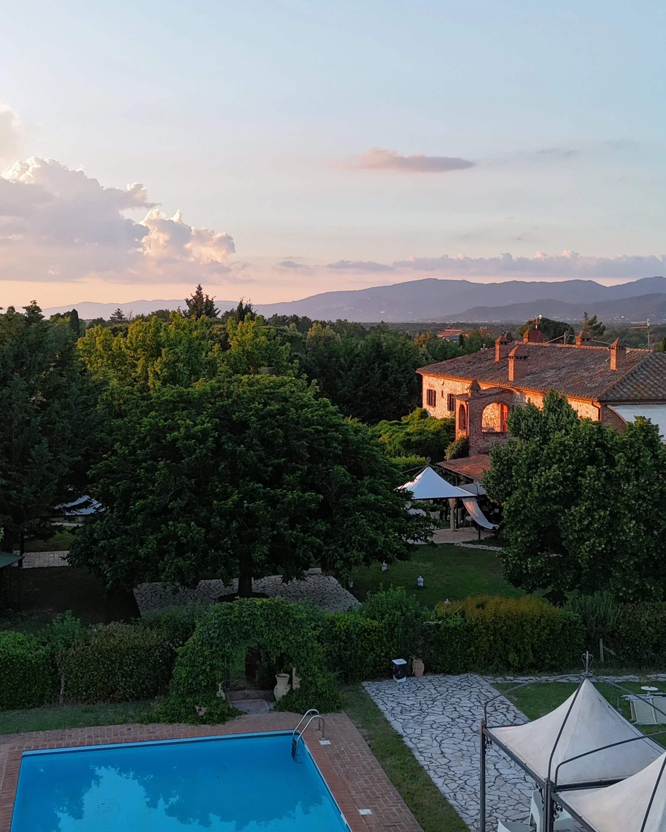 Vista di una piscina all'aperto, circondata da un giardino con alberi e arbusti, e un'area con sedie e gazebo. Sullo sfondo si vedono colline e montagne sotto un cielo con nuvole.