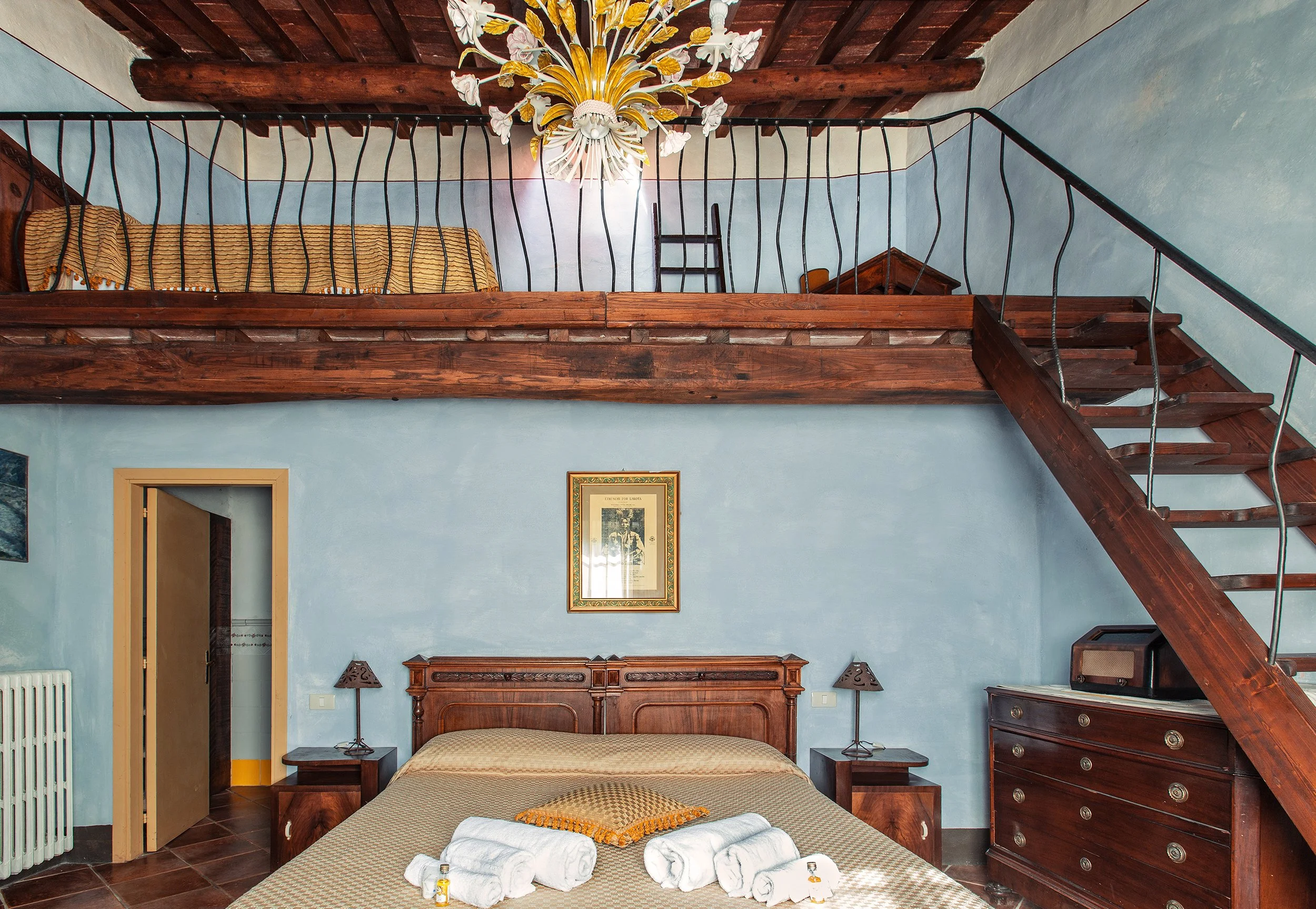 Camera da letto con pareti azzurre, letto con coperta beige, due comodini con lampade a forma di lanterna, quadro sulla parete, soffitto con travi di legno e lampadario decorativo, scala di legno che conduce a una soffitta.