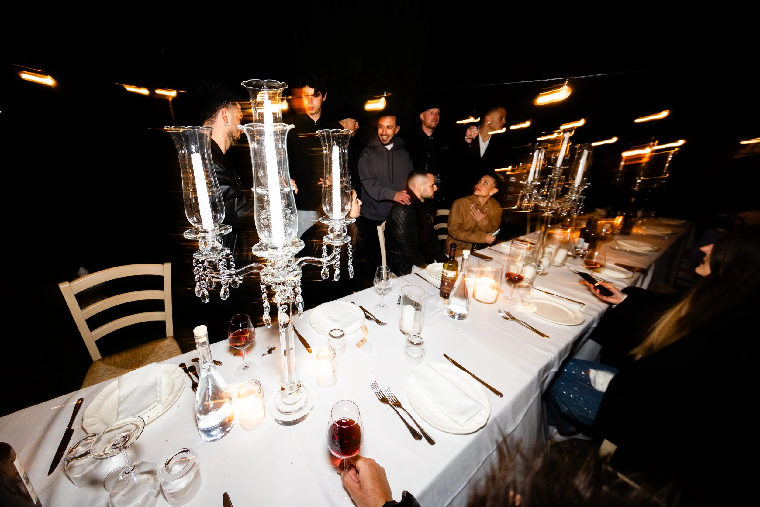 Persone che celebrano una cena di gruppo in un ambiente elegante con candelieri di cristallo e bicchieri di vino rosso, atmosfera calda e conviviale.