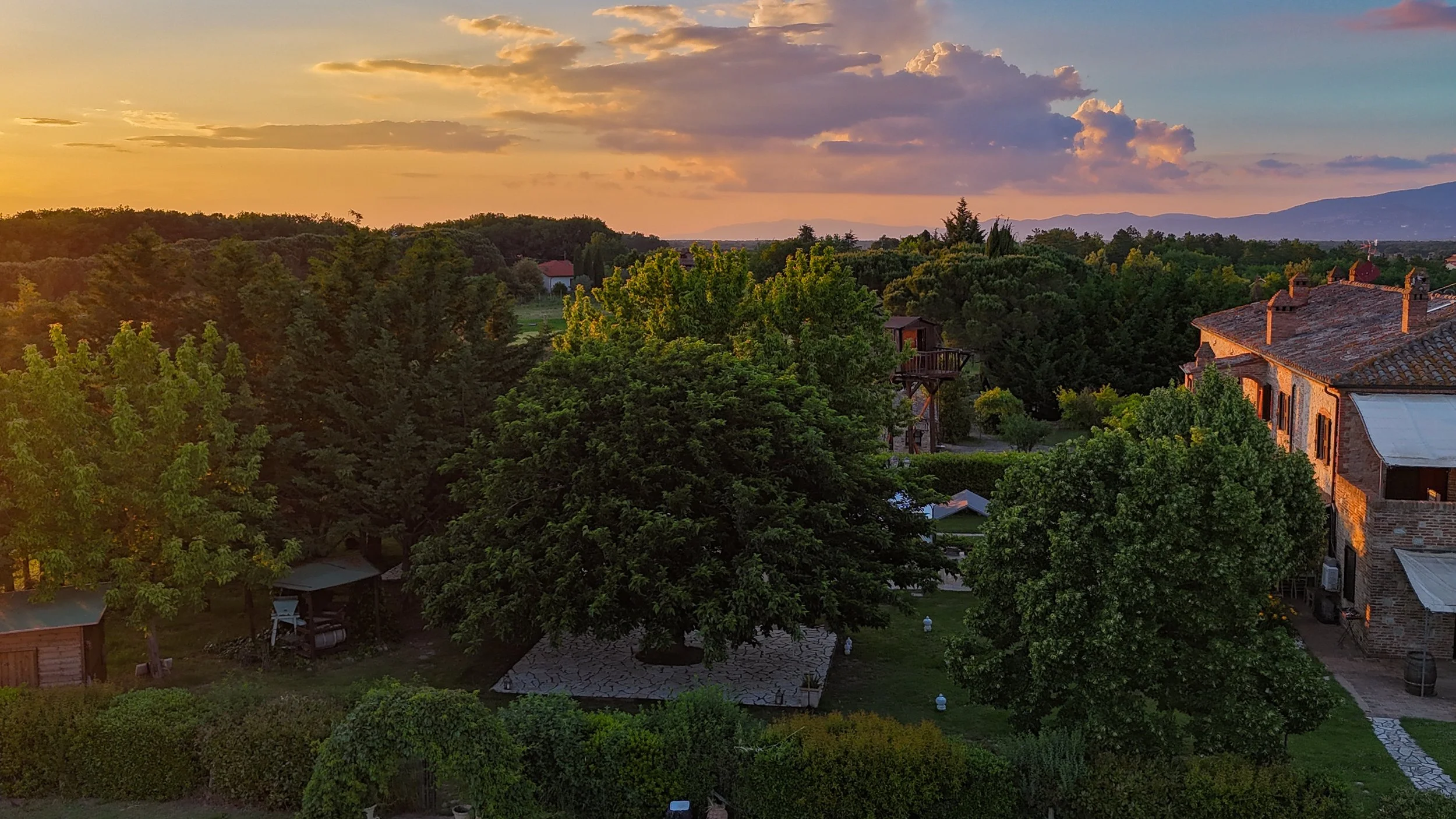 Paesaggio rurale al tramonto con alberi verdi, case in pietra con tetti di tegole, e un cielo colorato con nuvole soffici.