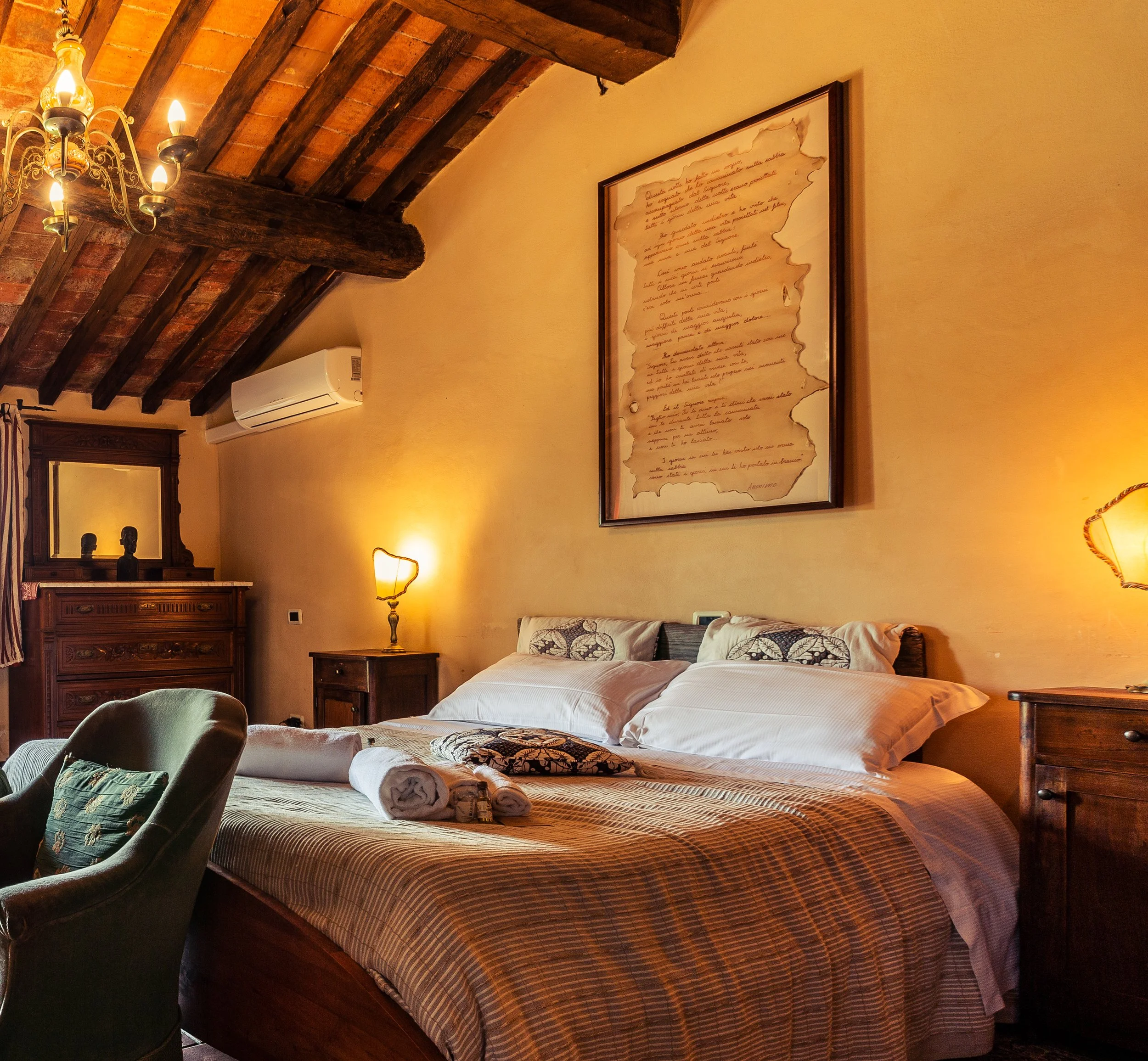 camera da letto con pareti beige, letto matrimoniale con coperte a quadretti, due asciugamani arrotolati e prodotti per il bagno, lampada da tavolo, quadro con scrittura a mano, parete con travi in legno e lampadari con luci calde, aria condizionata.
