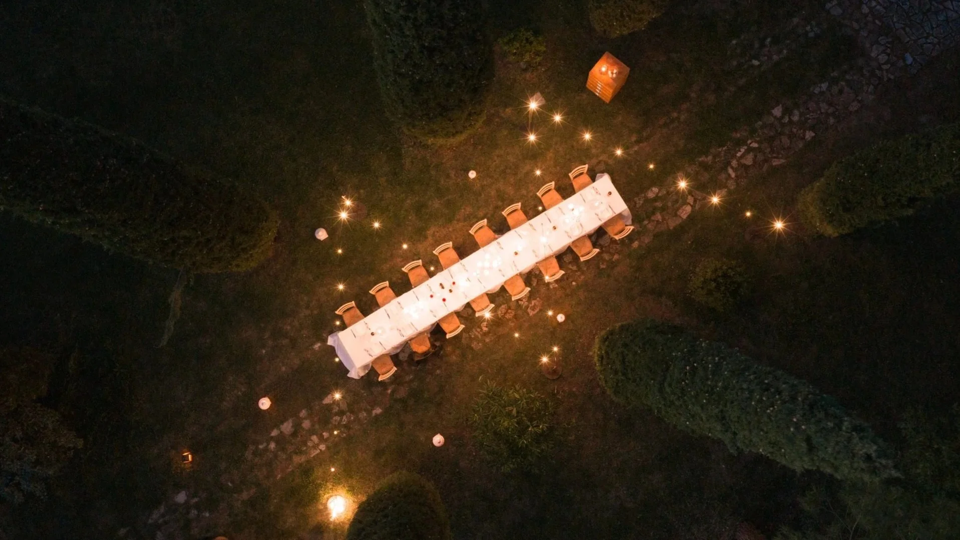 Vista dall'alto di un tavolo lungo con sedie intorno, illuminato da luci calde, in un giardino con siepi e pietre.