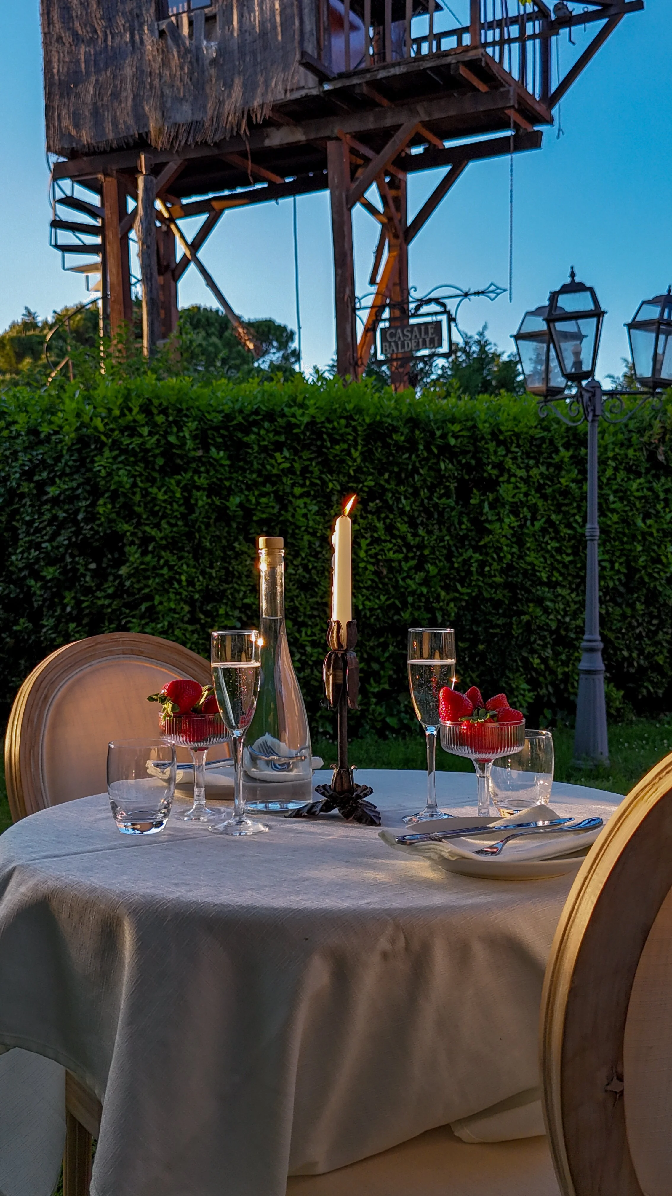 Tavola apparecchiata con bicchieri di prosecco, fragole in coppette, un mazzo di fiori e una candela, con un sfondo di luci e una struttura in legno all'aperto.