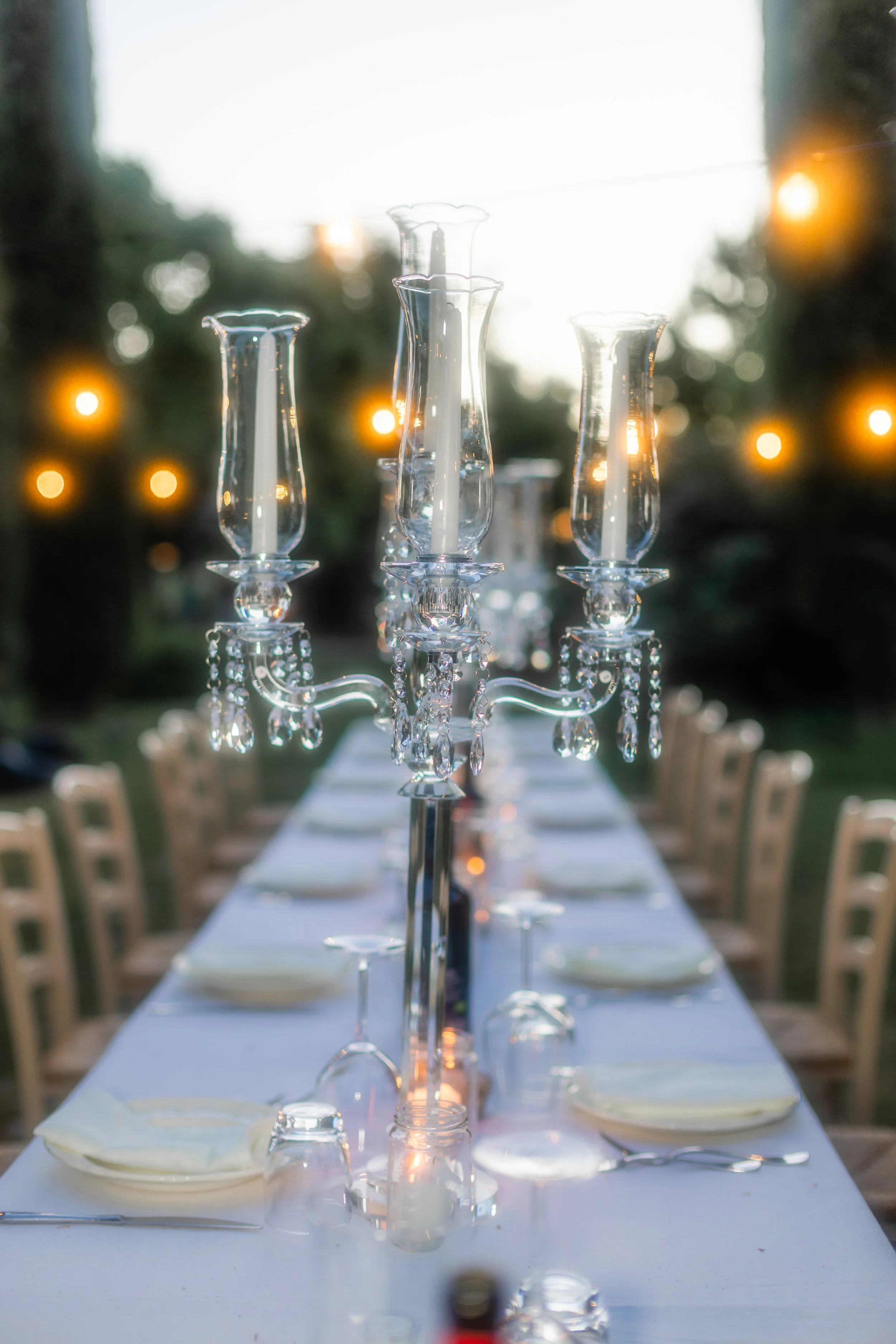 Una lunga tavola decorata con un centrotavola composto da candelabri di cristallo con candele accese. Sul tavolo ci sono piatti, posate e tovaglioli, pronti per un pasto. La scena è all'aperto durante il crepuscolo, con luci calde e sfocate sullo sfondo.