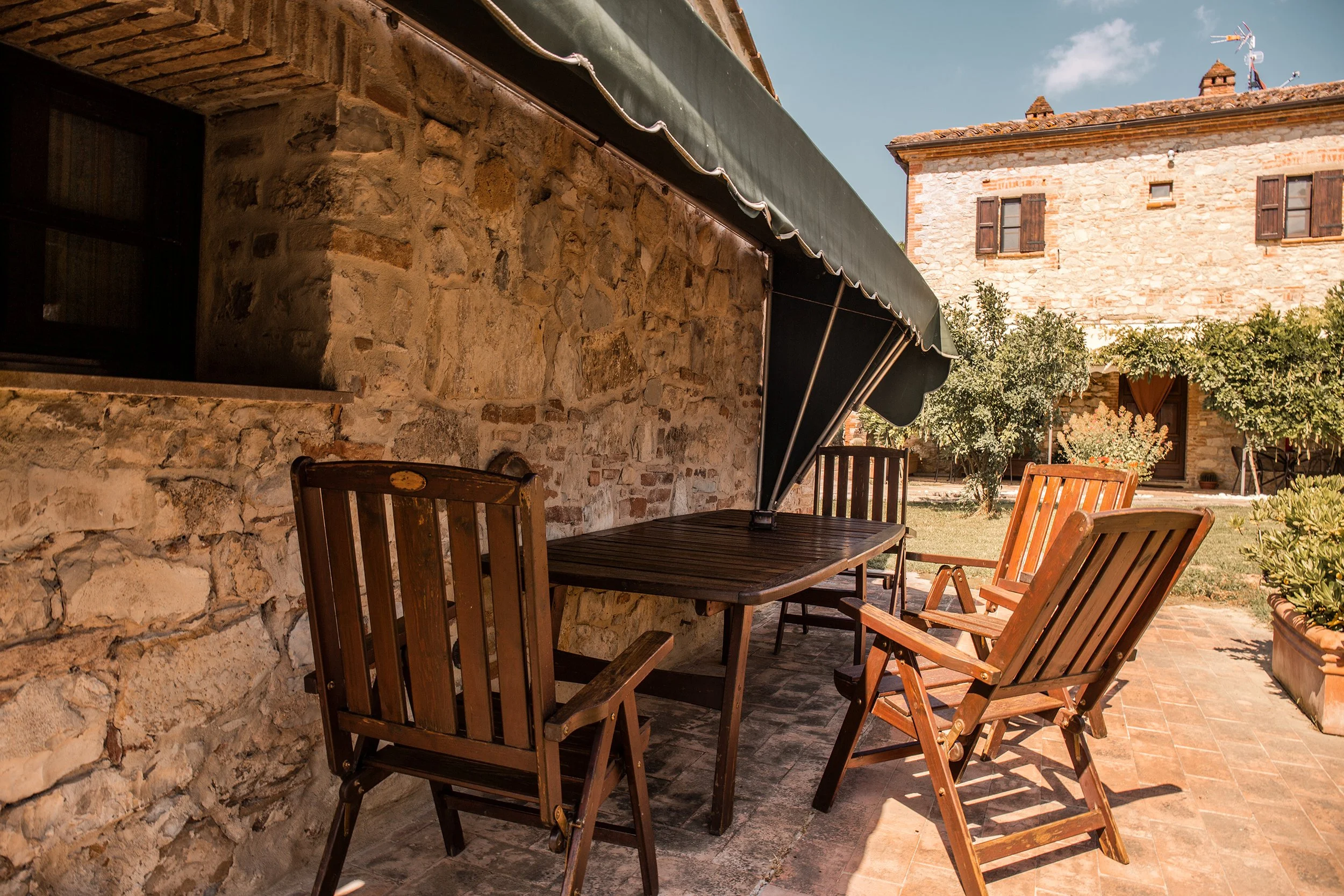 Zona esterna di una casa rustica con tavolo e sedie di legno, sembra essere un patio o un giardino con piante e un edificio in pietra con finestre in legno.