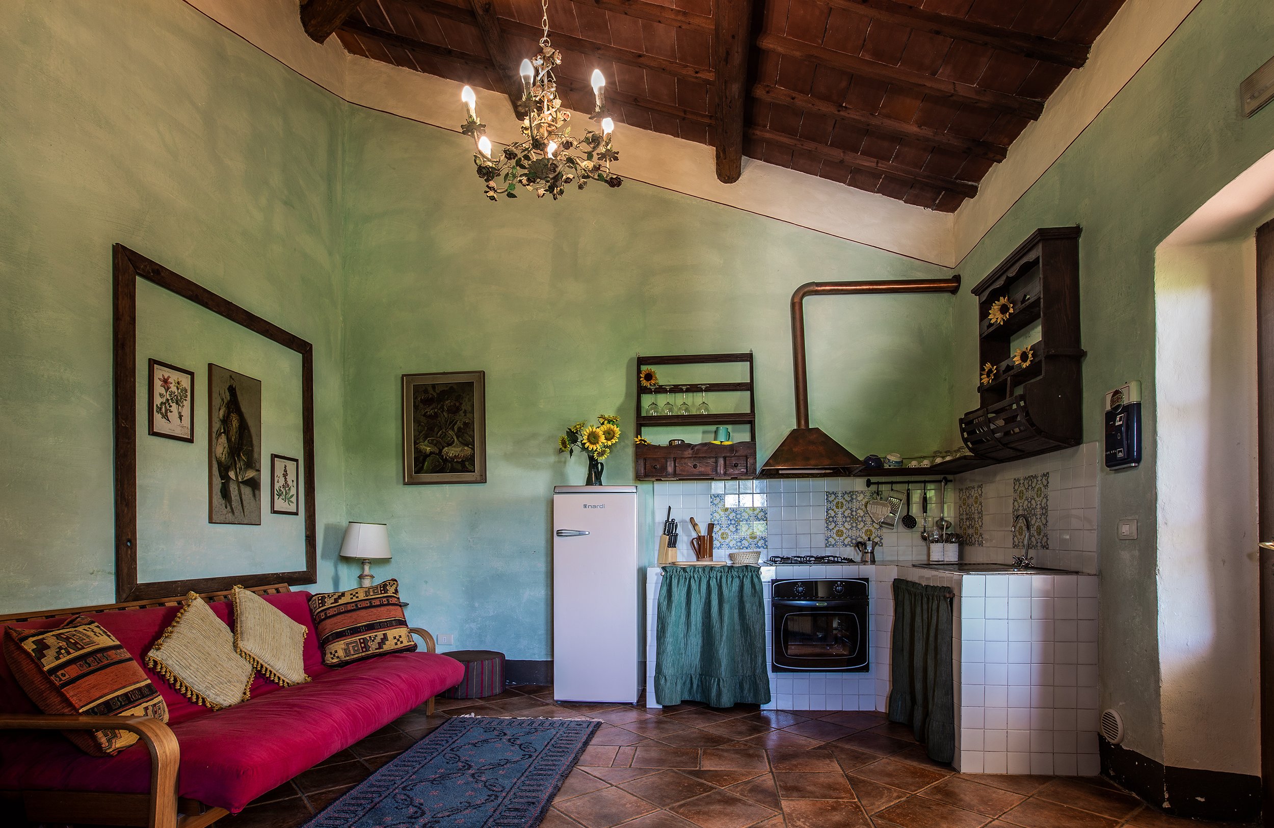 Interno di una cucina con pareti verdi, soffitto in legno con travi visibili, lampadario decorato con fiori, cucina con stoviglie e frigorifero, divano con cuscini a motivi geometrici