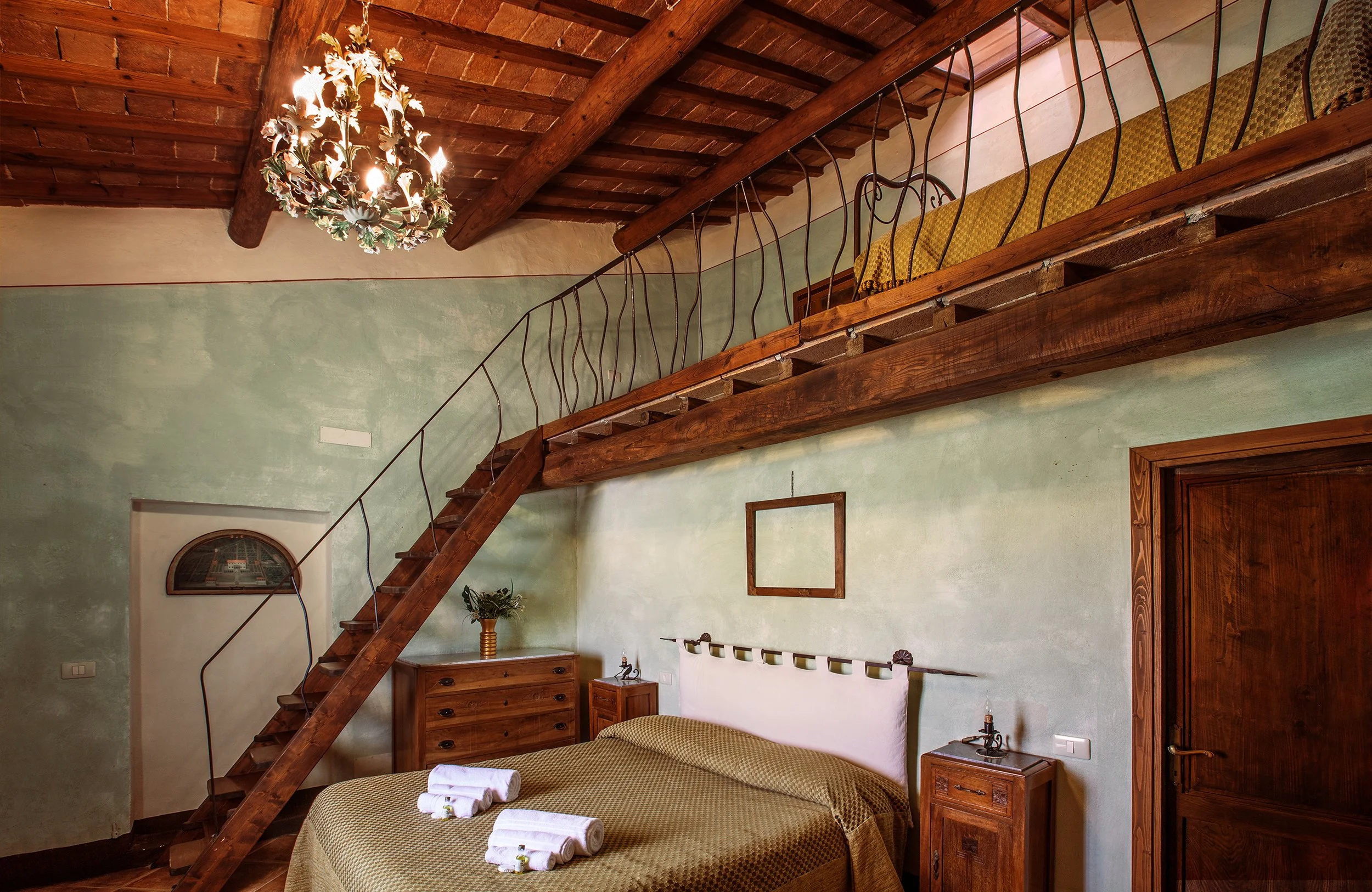 Camera da letto con soffitto in legno, letto con lenzuola verdi, due asciugamani arrotolati sul letto, lampadario decorativo, pareti verdi, mobile in legno, decorazioni in stanza.