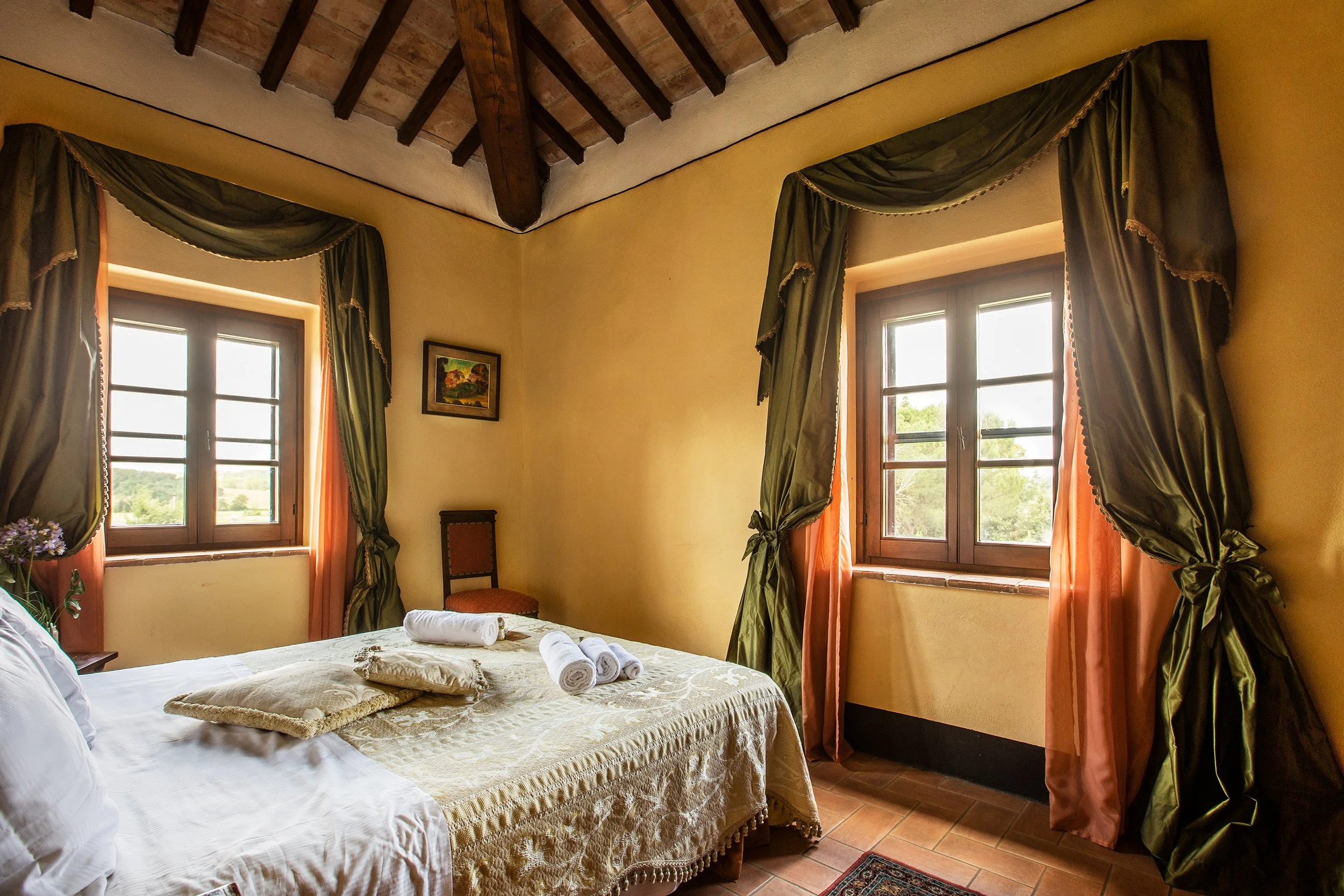 Camera da letto con due finestre, tende verdi con nappe, letto con biancheria bianca e asciugamani arrotolati, pareti gialle e soffitto con travi in legno.