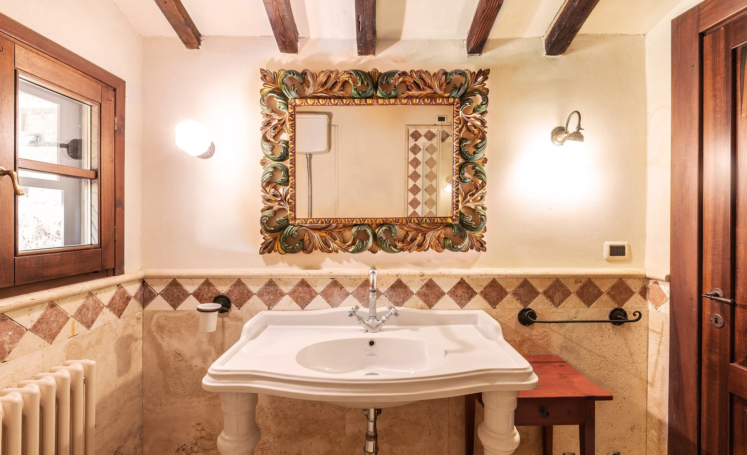 Specchio decorativo sopra un lavandino con due gambe, in un bagno con pareti color avorio, lampade a muro e finestra in legno.