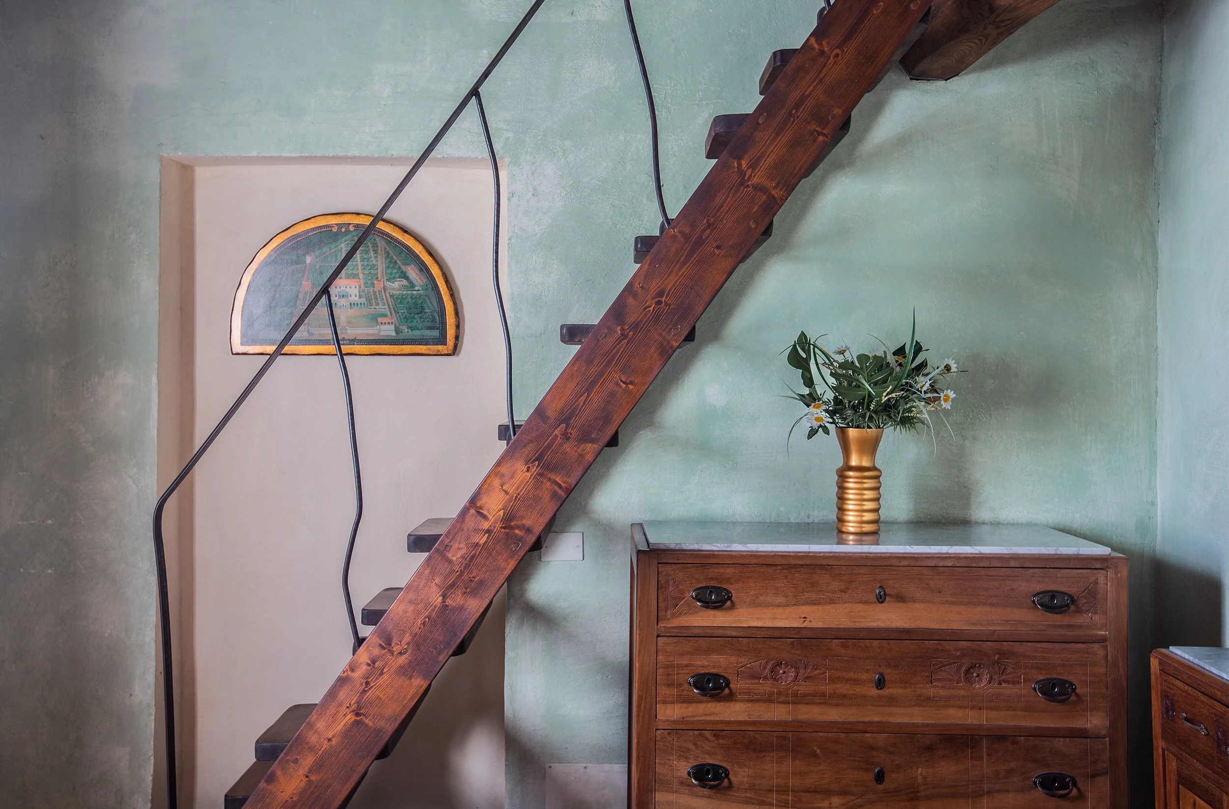 Scala in legno con ringhiera in ferro nero, un mobile in legno con quattro cassetti, un vaso dorato con fiori e piante verdi, pareti verde pastello con decorazioni.