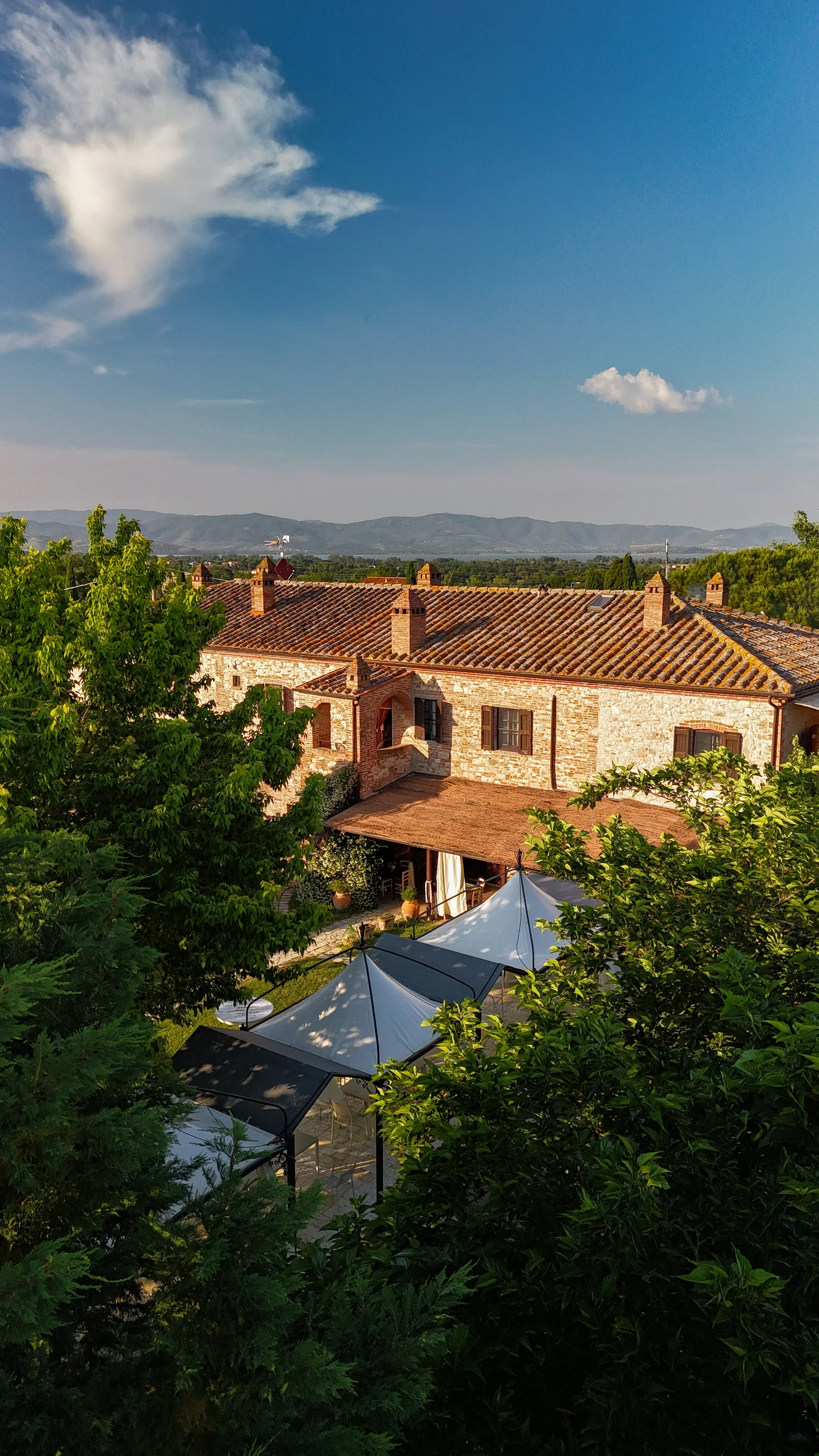 Veduta di un edificio in stile rustico con tetto in tegole, circondato da alberi verdi, con un cielo blu e nuvole sparse sullo sfondo.