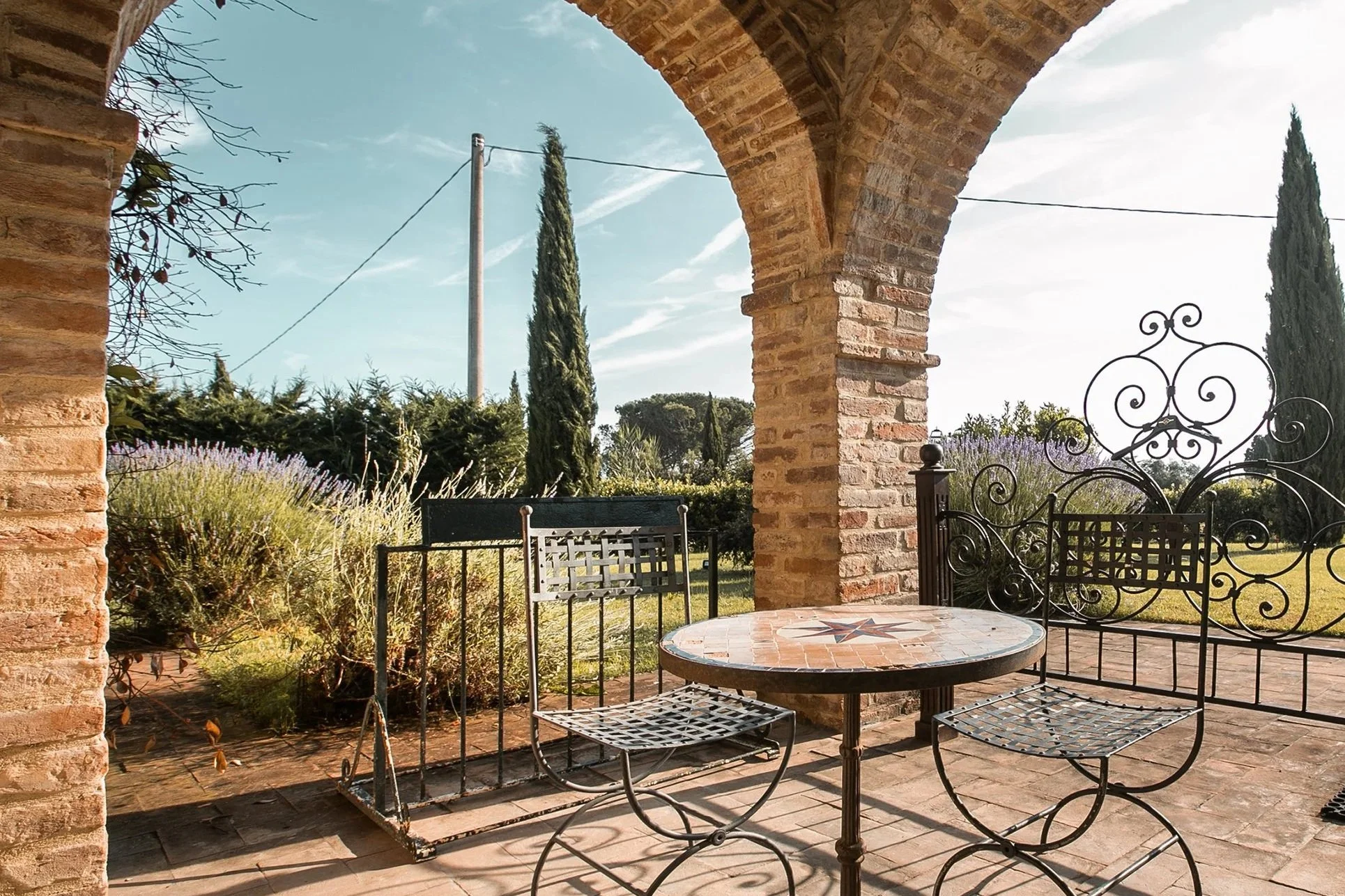 Una terrazza con tavolo rotondo in pietra decorata, sedie di ferro nero, una parete di mattoni e un arco di mattoni, con piante e alberi sullo sfondo e un cielo blu.
