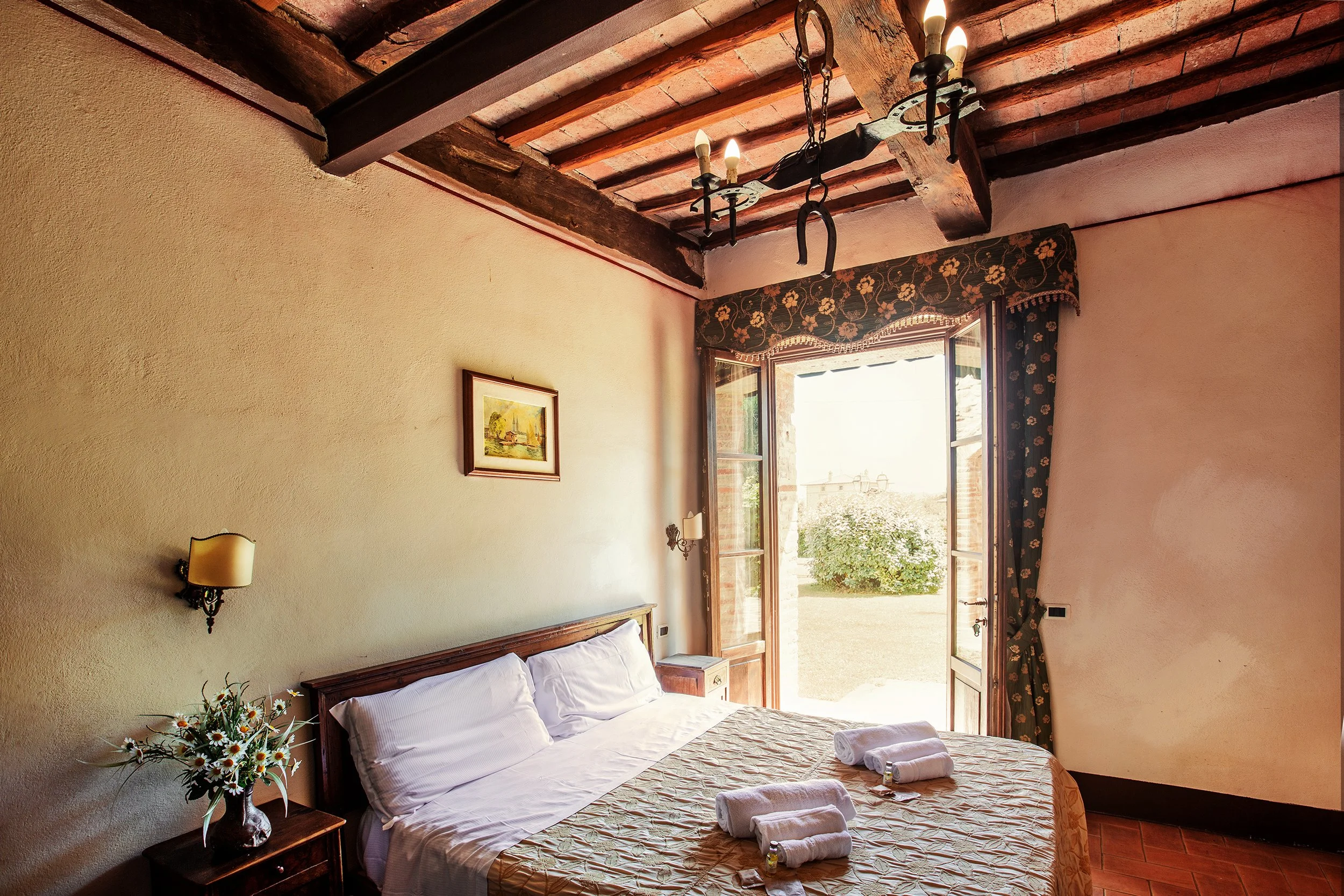 Camera da letto con letto matrimoniale, coperte e asciugamani arrotolati, finestra aperta con tendaggi a fiori e vista su un giardino, pareti di colore beige e soffitto con travi di legno e lampadario