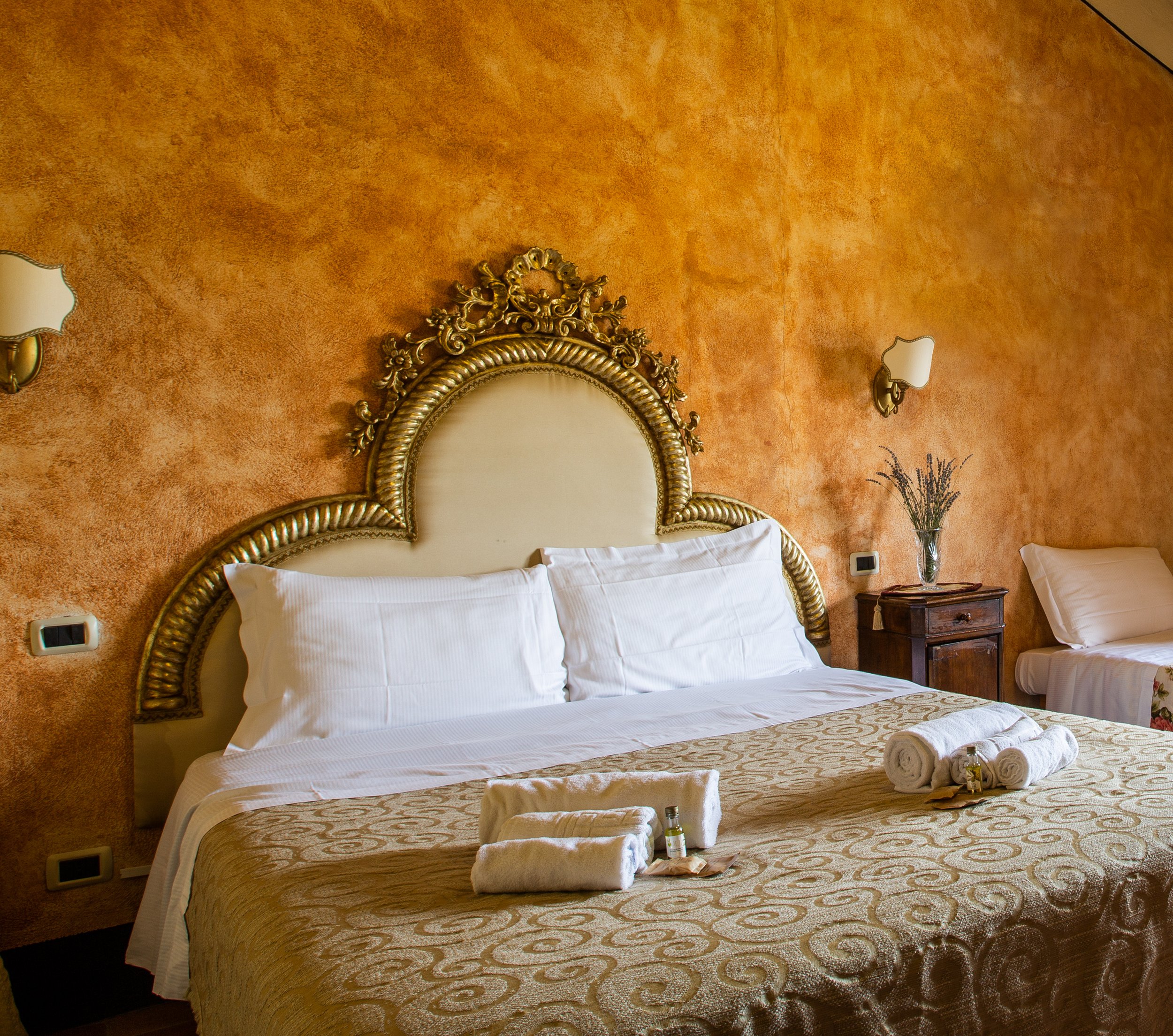 Letto matrimoniale con lenzuola bianche, due asciugamani arrotolati e alcune bottiglie di olio essenziale sulla coperta, lampade ai lati del letto, specchio dorato sopra la testata, pareti arancioni con decorazioni, vaso con fiori sulla sinistra.
