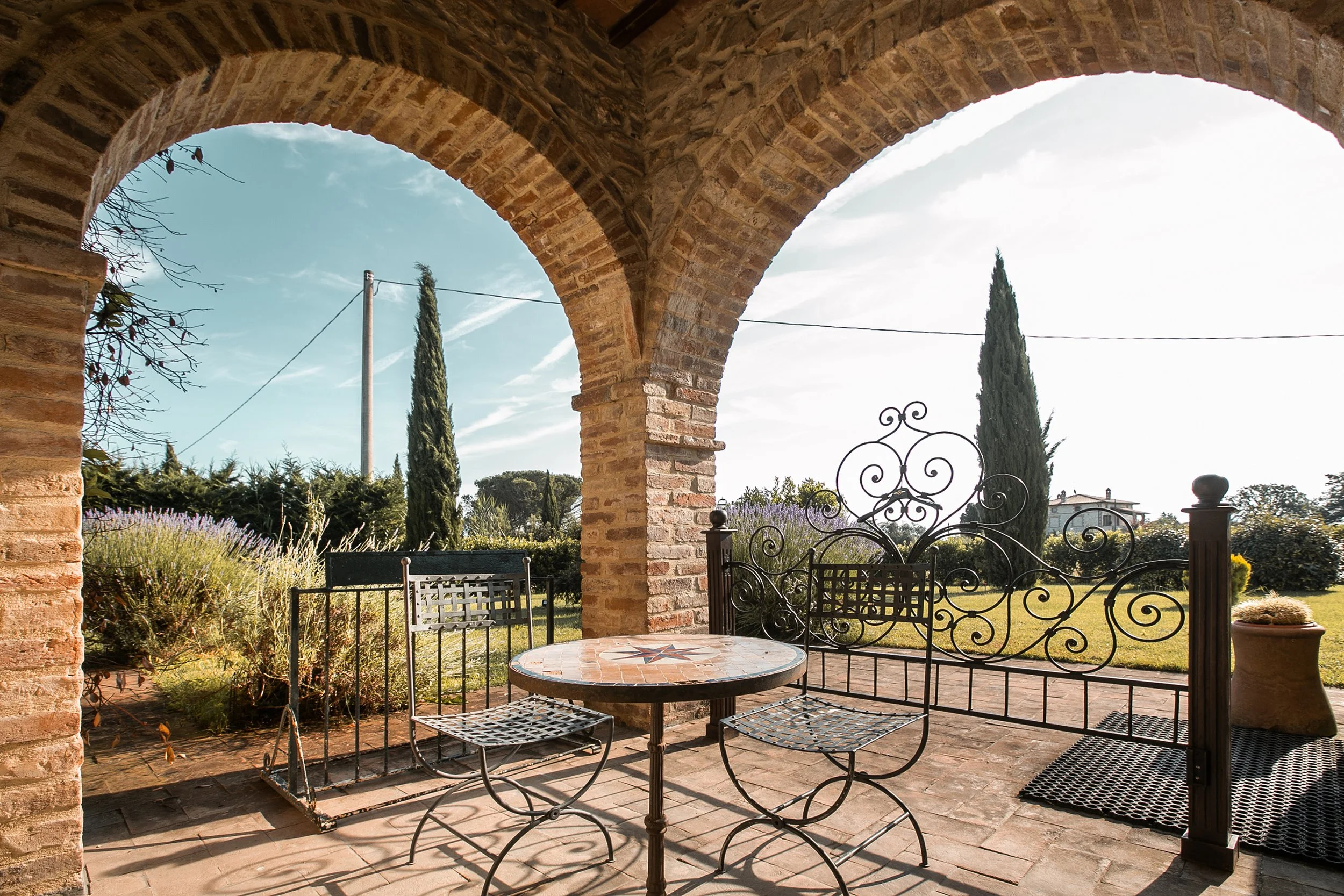 Veduta di un patio con archi in mattoni, tavolino decorato con mosaico, sedie in metallo, giardino con arbusti e alberi, e cielo azzurro con nuvole.
