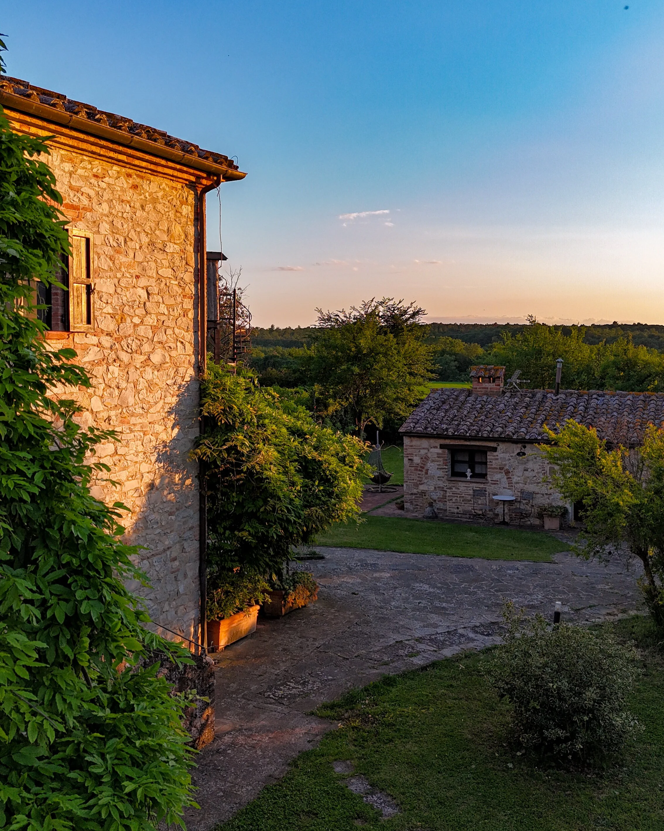 Una scena al tramonto con case in pietra, piante verdi e un cielo blu con alcune nuvole sparse, ambientata in campagna.
